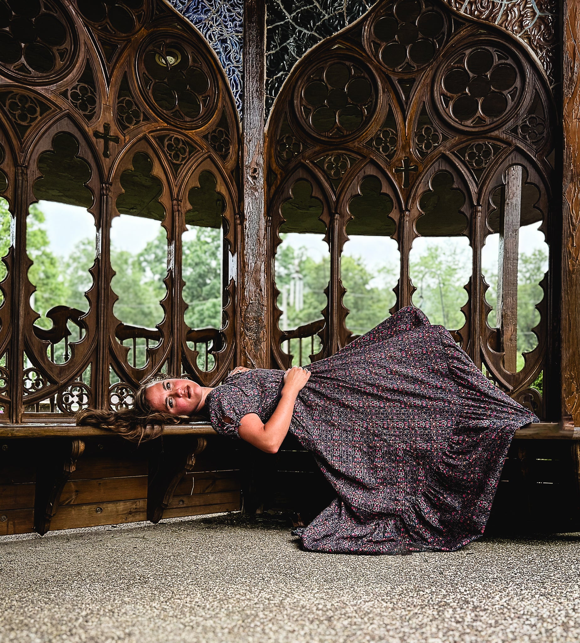 Woman in a dark modest nursing dress lying on a wooden bench with intricate carvings, surrounded by greenery.