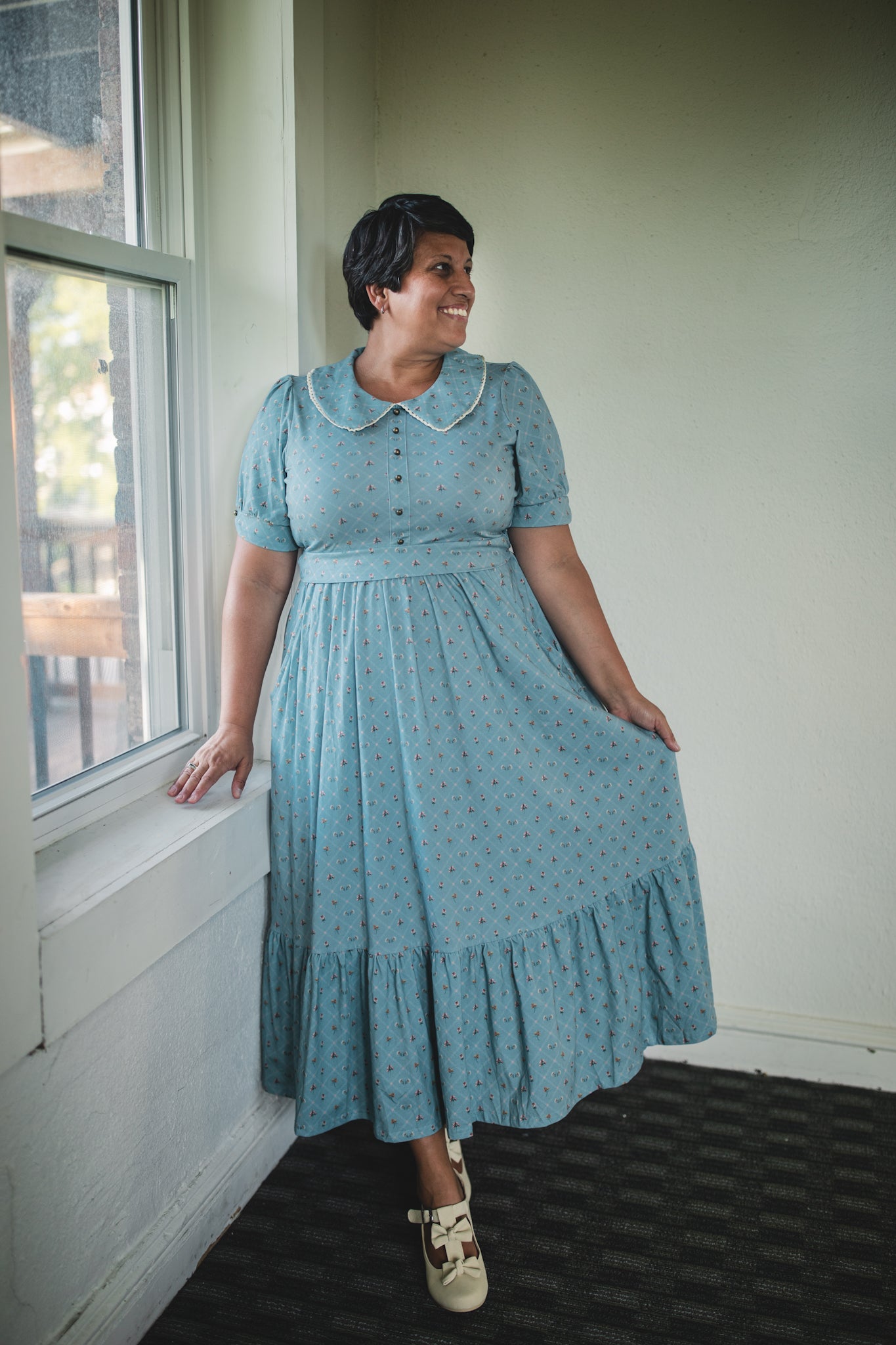 Woman wearing a blue modest nursing dress standing by a window indoors.