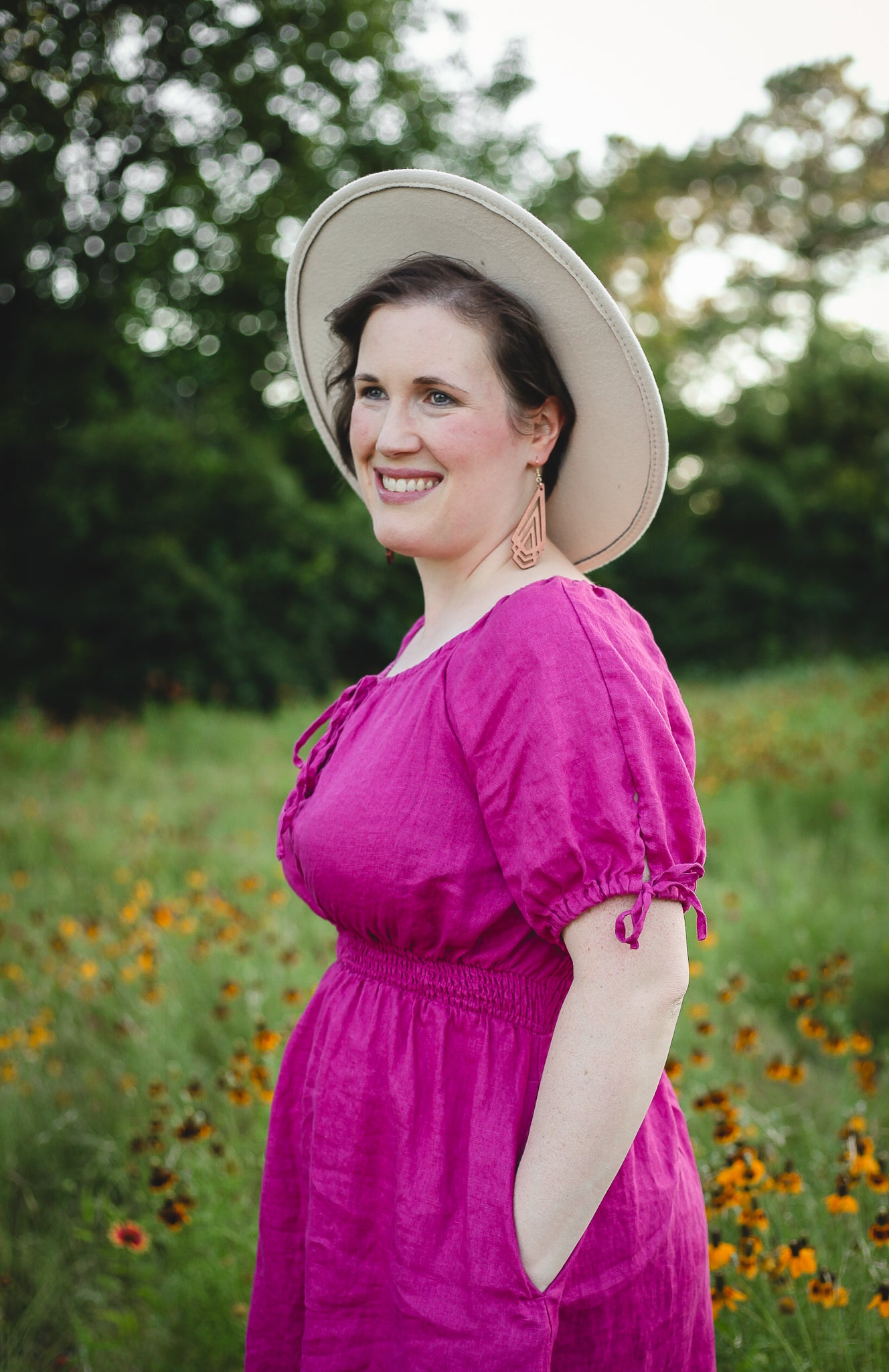 Woman in a pink modest nursing dress and white hat standing in a field with greenery and flowers.