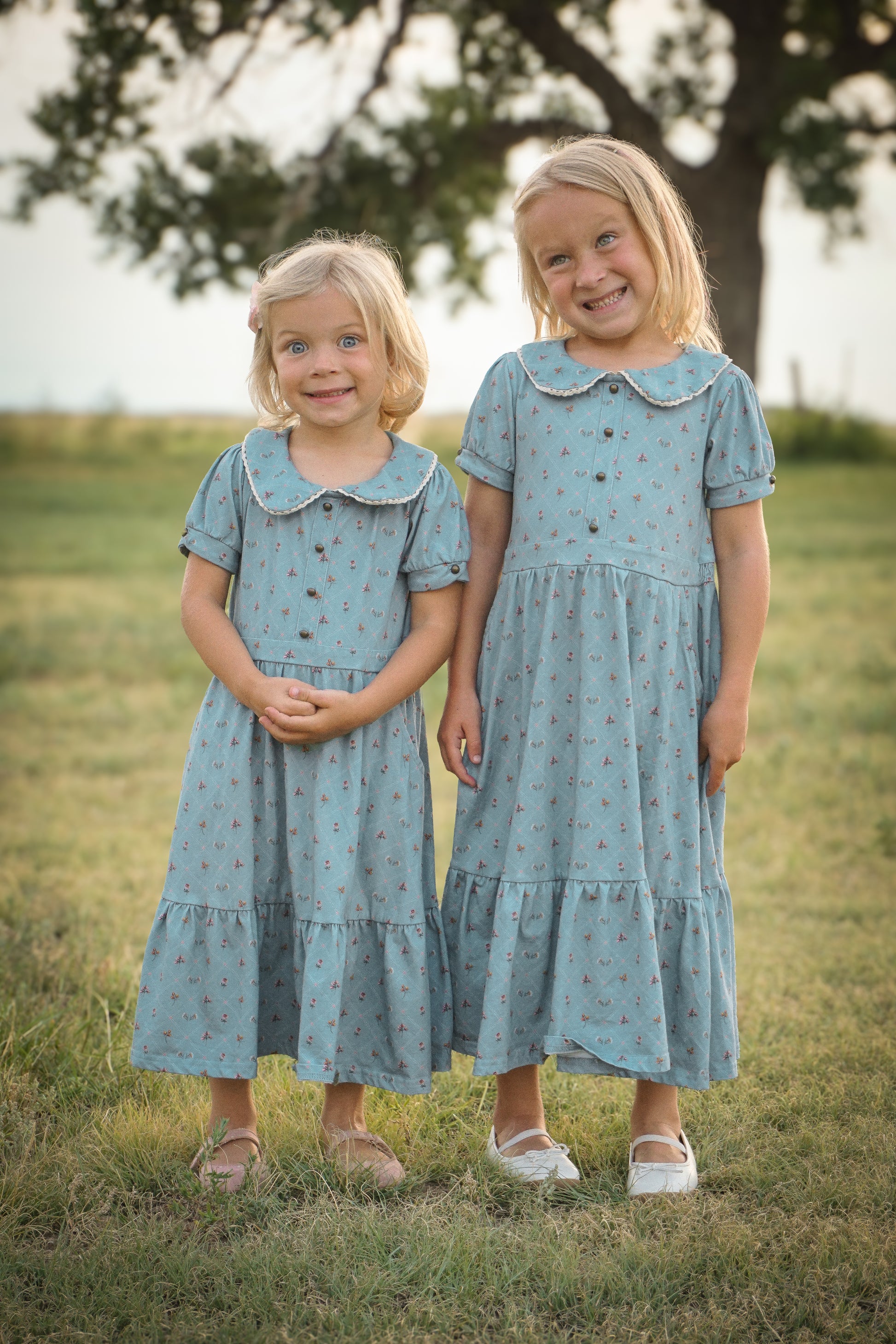 Two young girls in matching blue dresses standing in a grassy field.