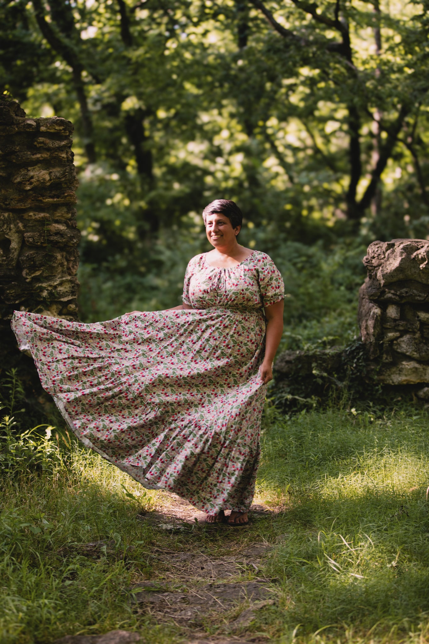 Woman in a floral modest nursing dress standing in a forested area with sunlight filtering through the trees.