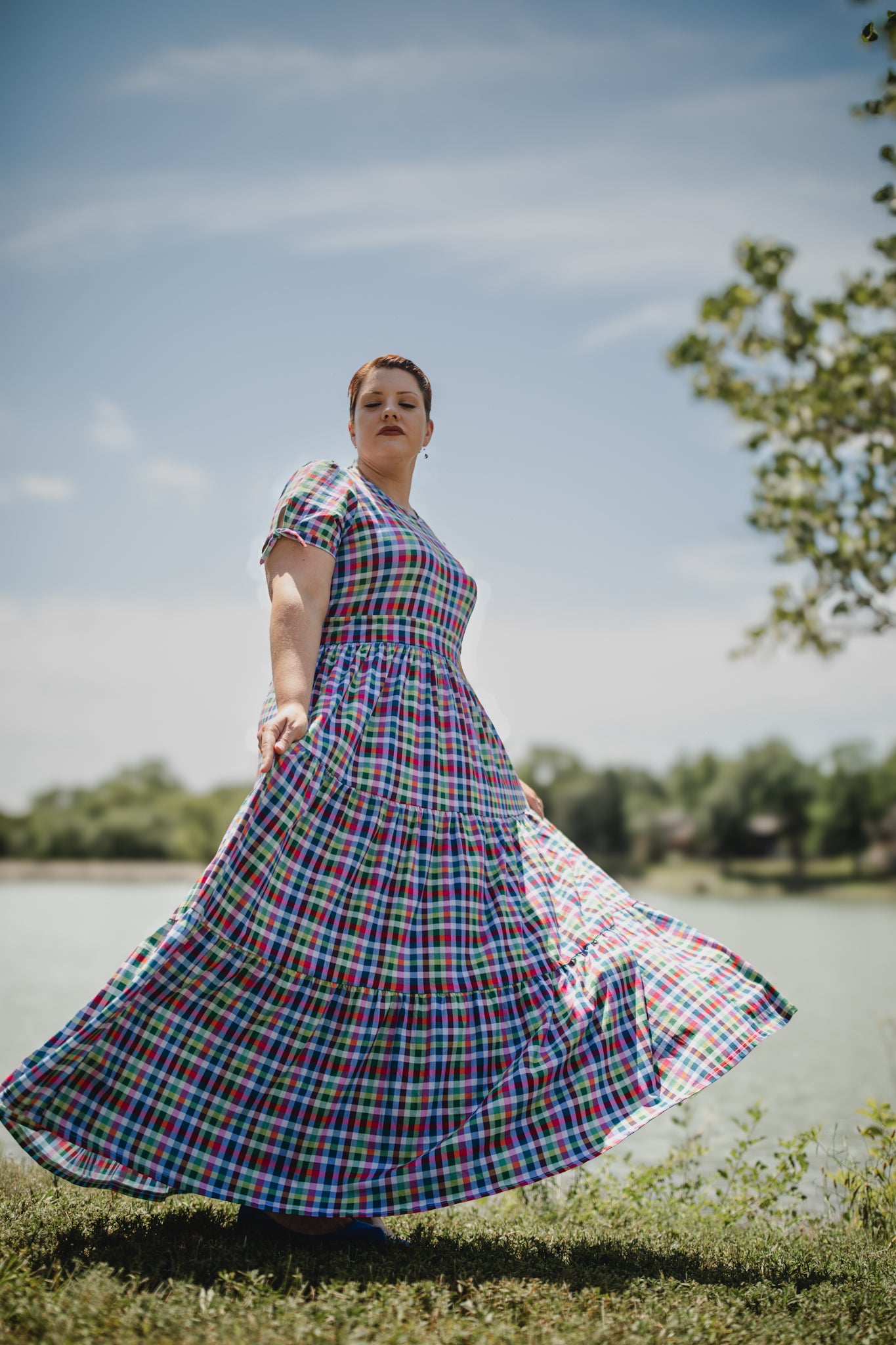 Woman in a plaid modest nursing dress standing outdoors with a clear sky and trees in the background