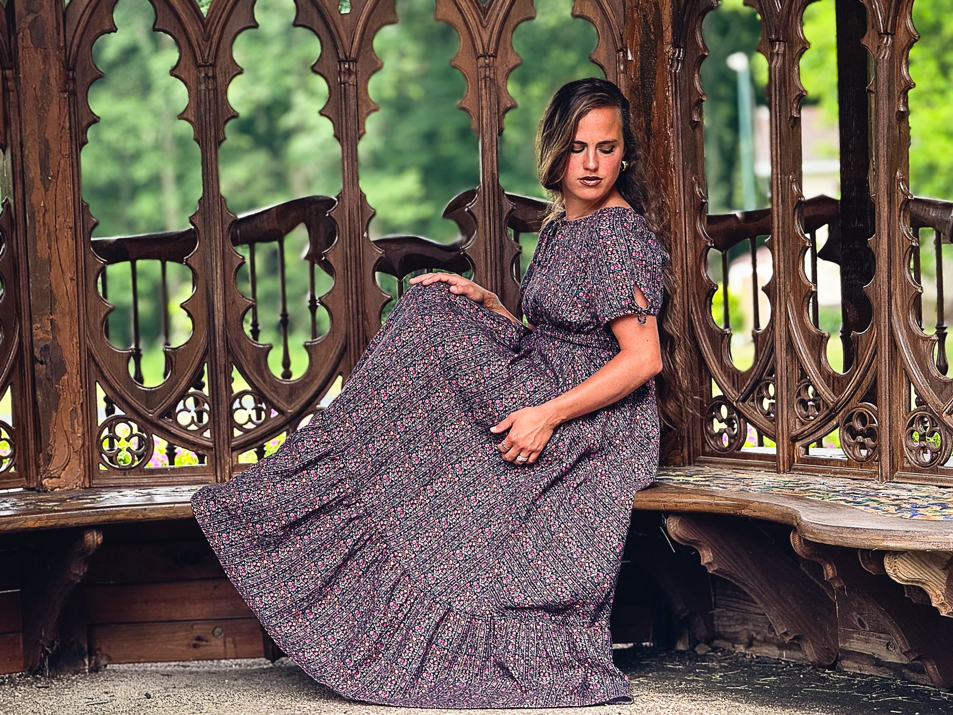 Woman in a floral modest nursing dress sitting on an ornate wooden bench with a blurred green background