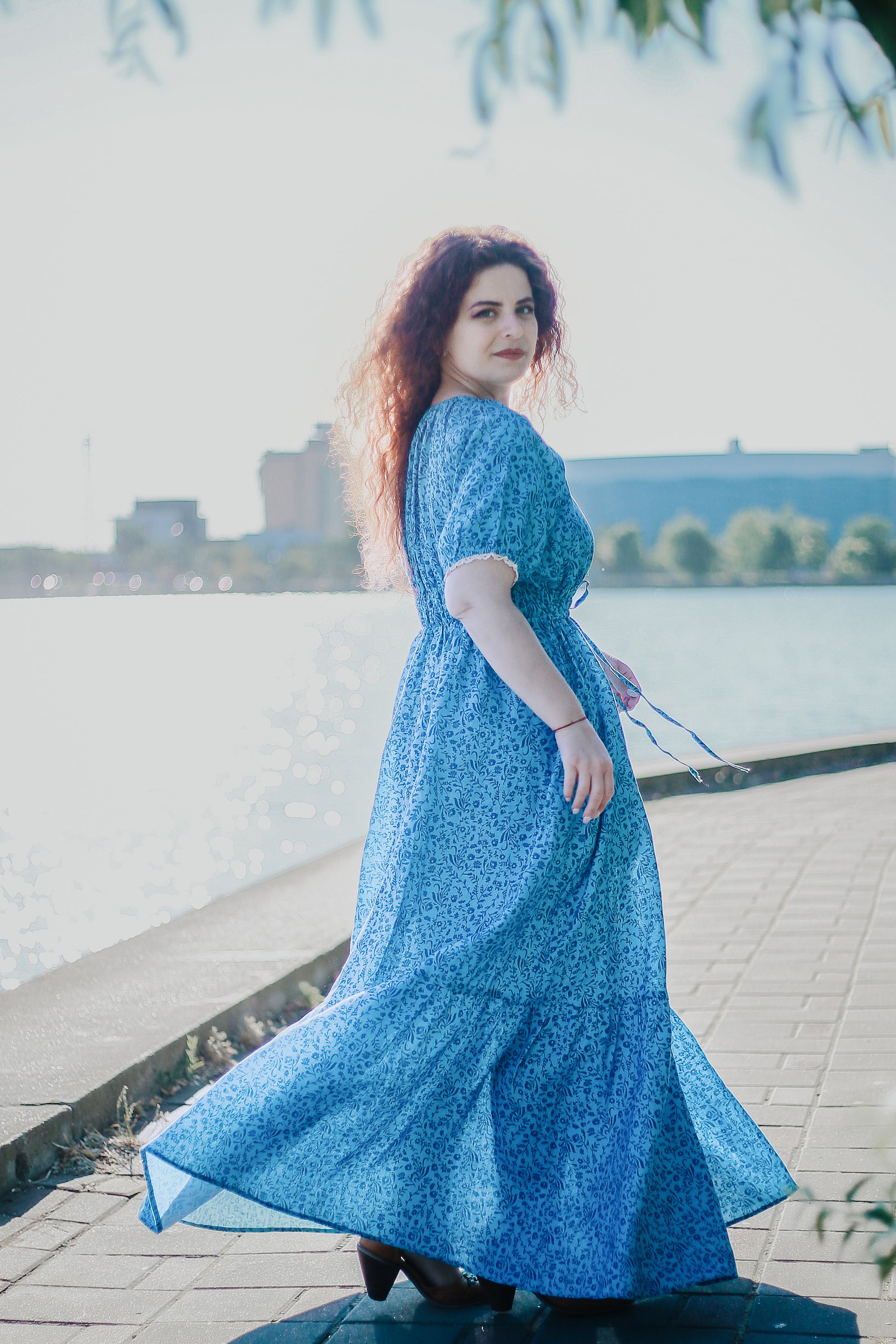 Woman in a blue modest nursing dress standing by a waterfront with buildings in the background