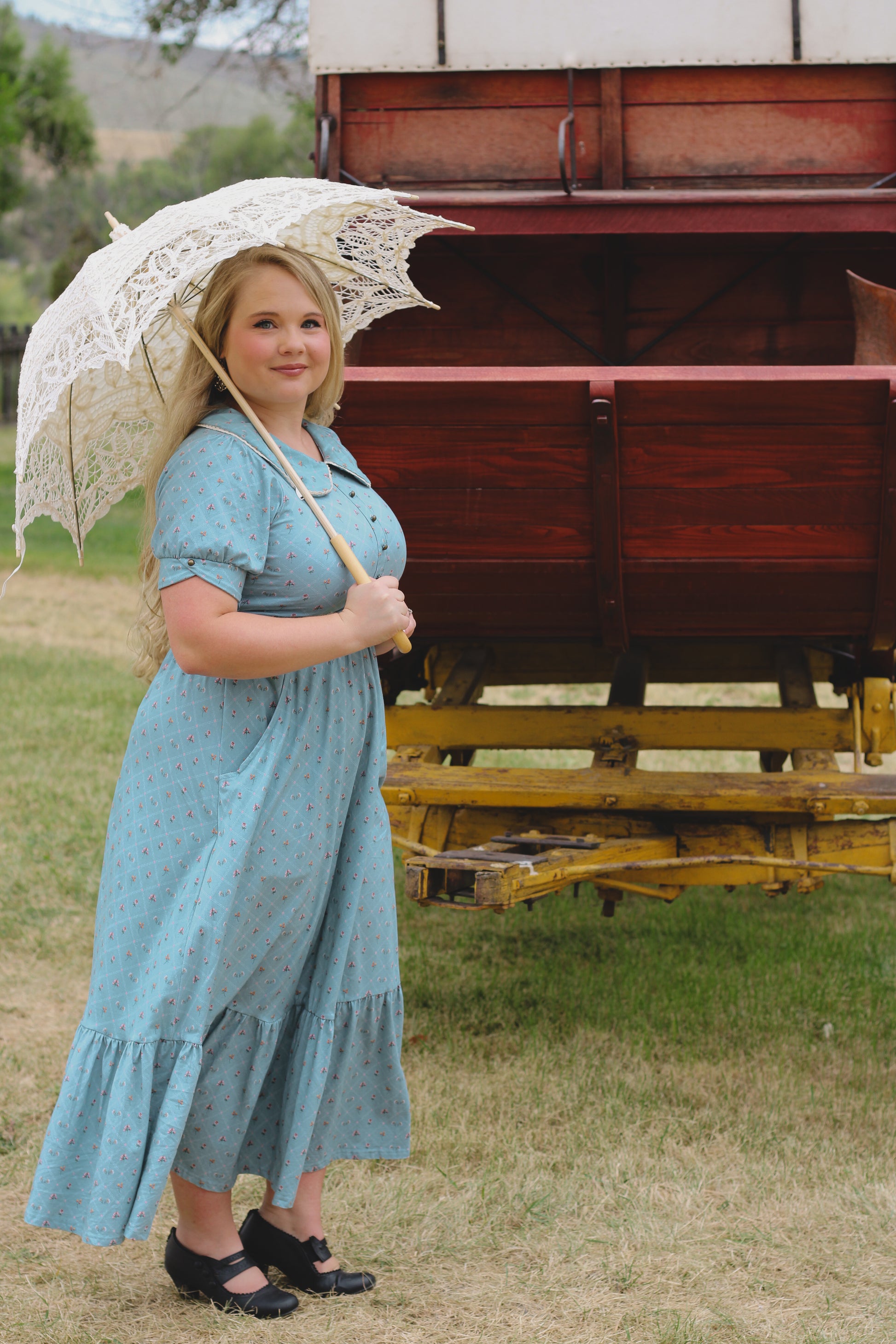 Woman in a blue modest nursing dress holding a lace parasol in front of a wooden cart.