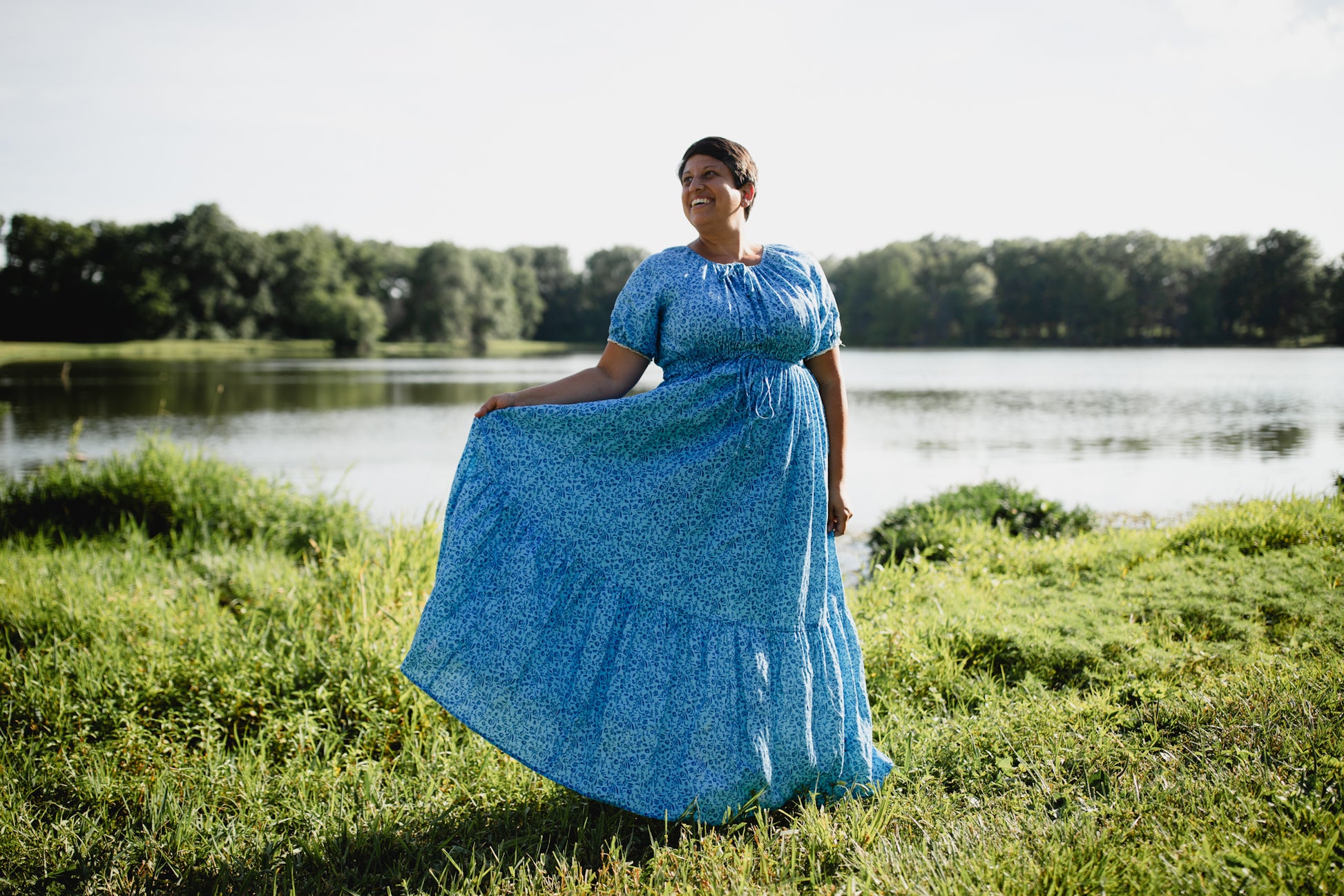 Woman in a blue modest nursing dress standing in a grassy area near a body of water with trees in the background.