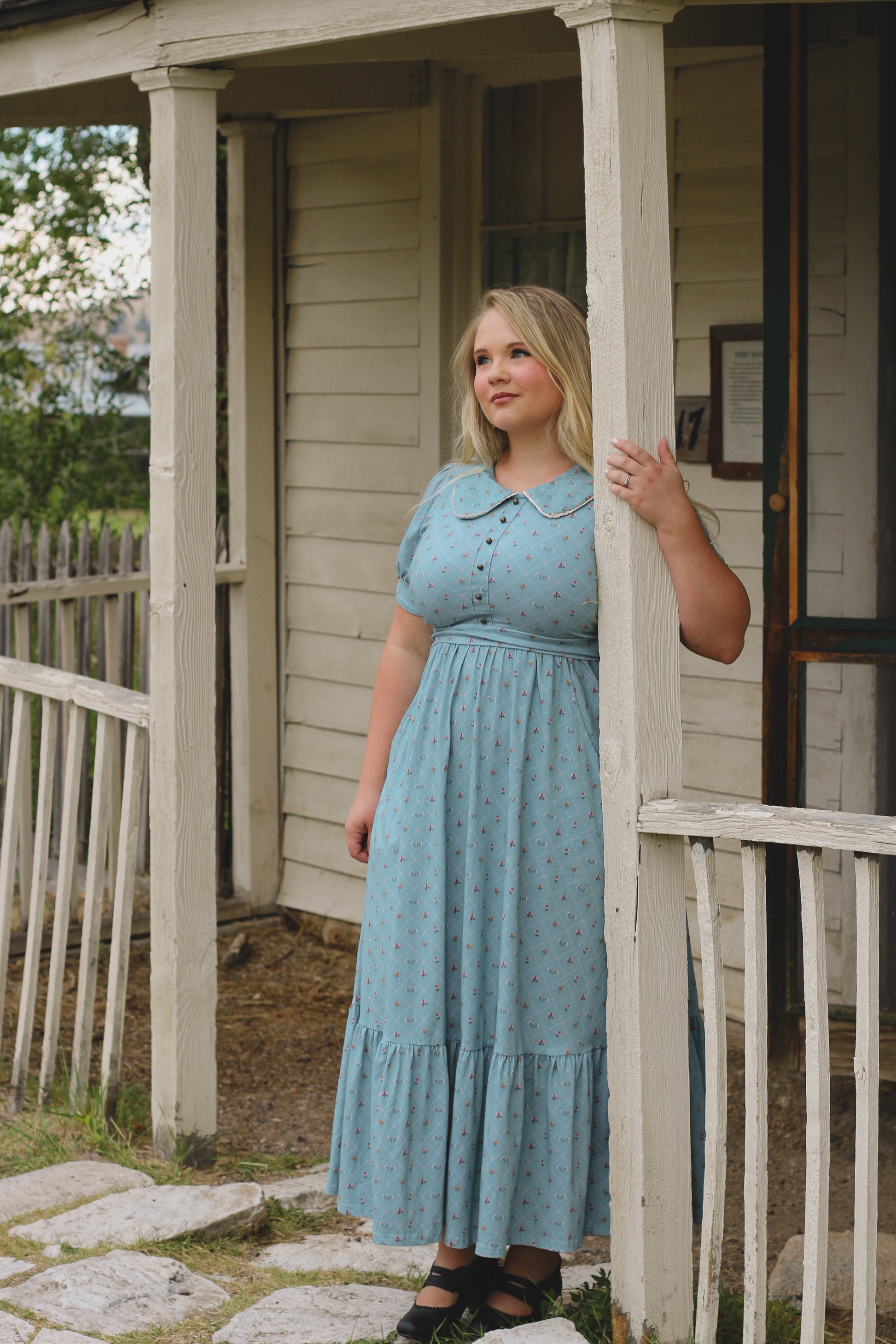 Woman in a blue modest nursing dress standing on a porch of a house.