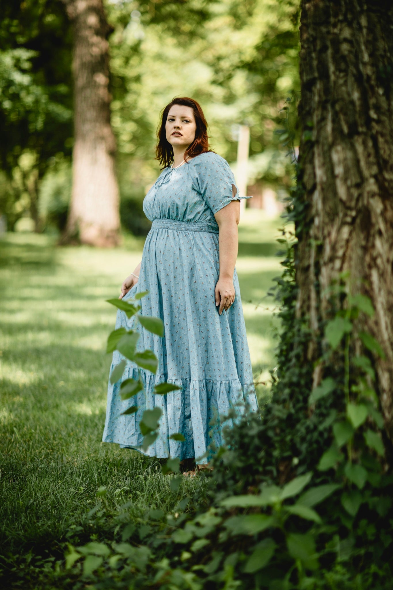 Woman in a light blue modest nursing dress standing in a park with trees and grass.