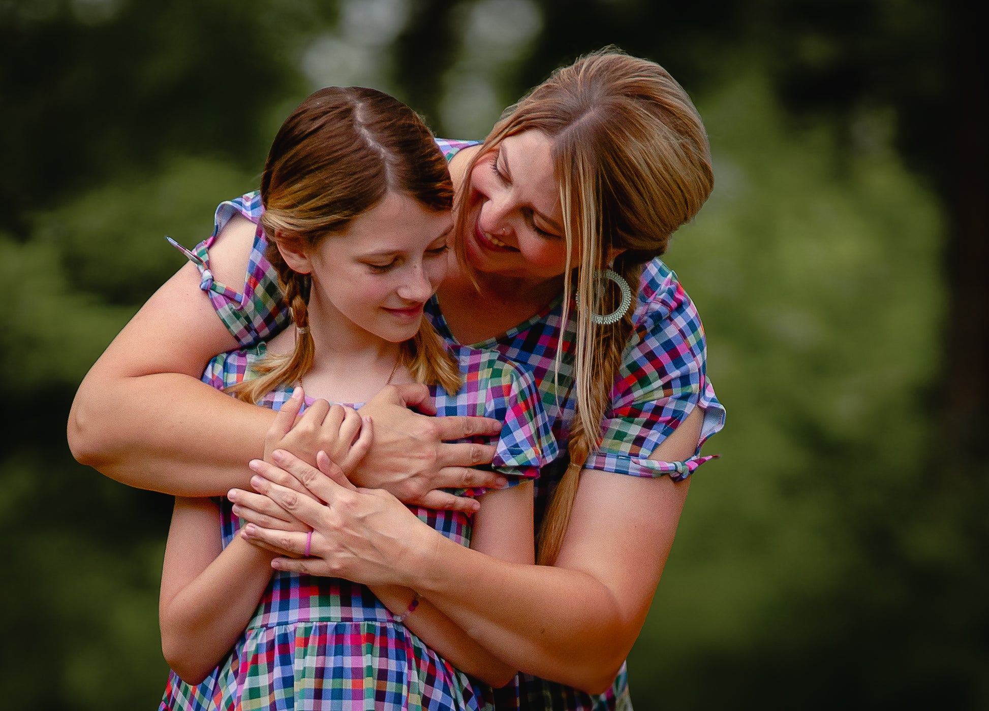 Woman and young girl in modest dresses hugging outdoors with a blurred green background