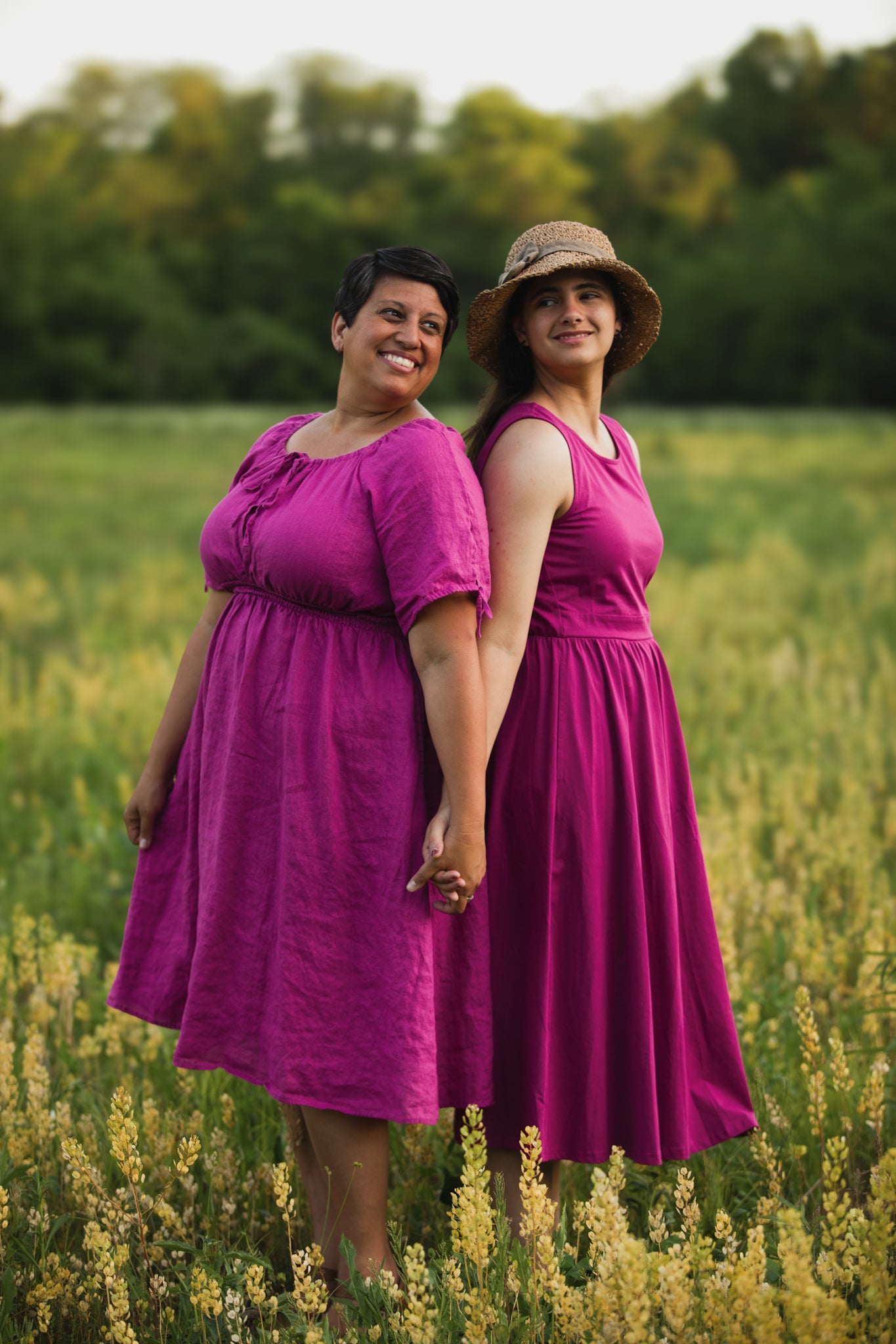 Two women in pink modest nursing dresses standing in a field with greenery in the background