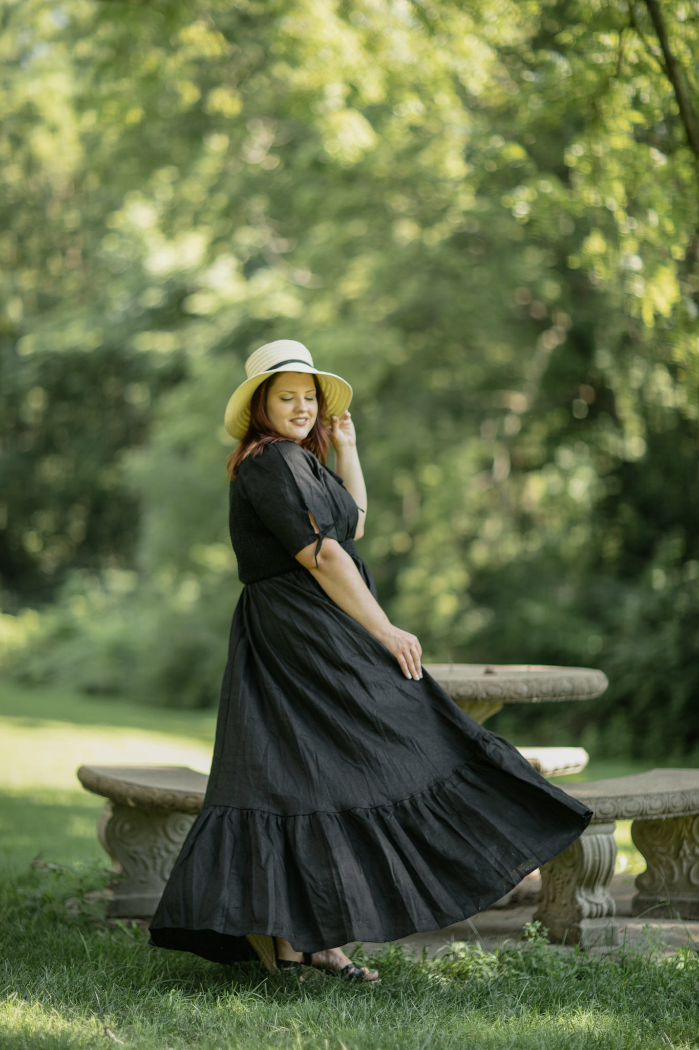 Woman in a black modest nursing dress and hat sitting on a stone bench in a park.