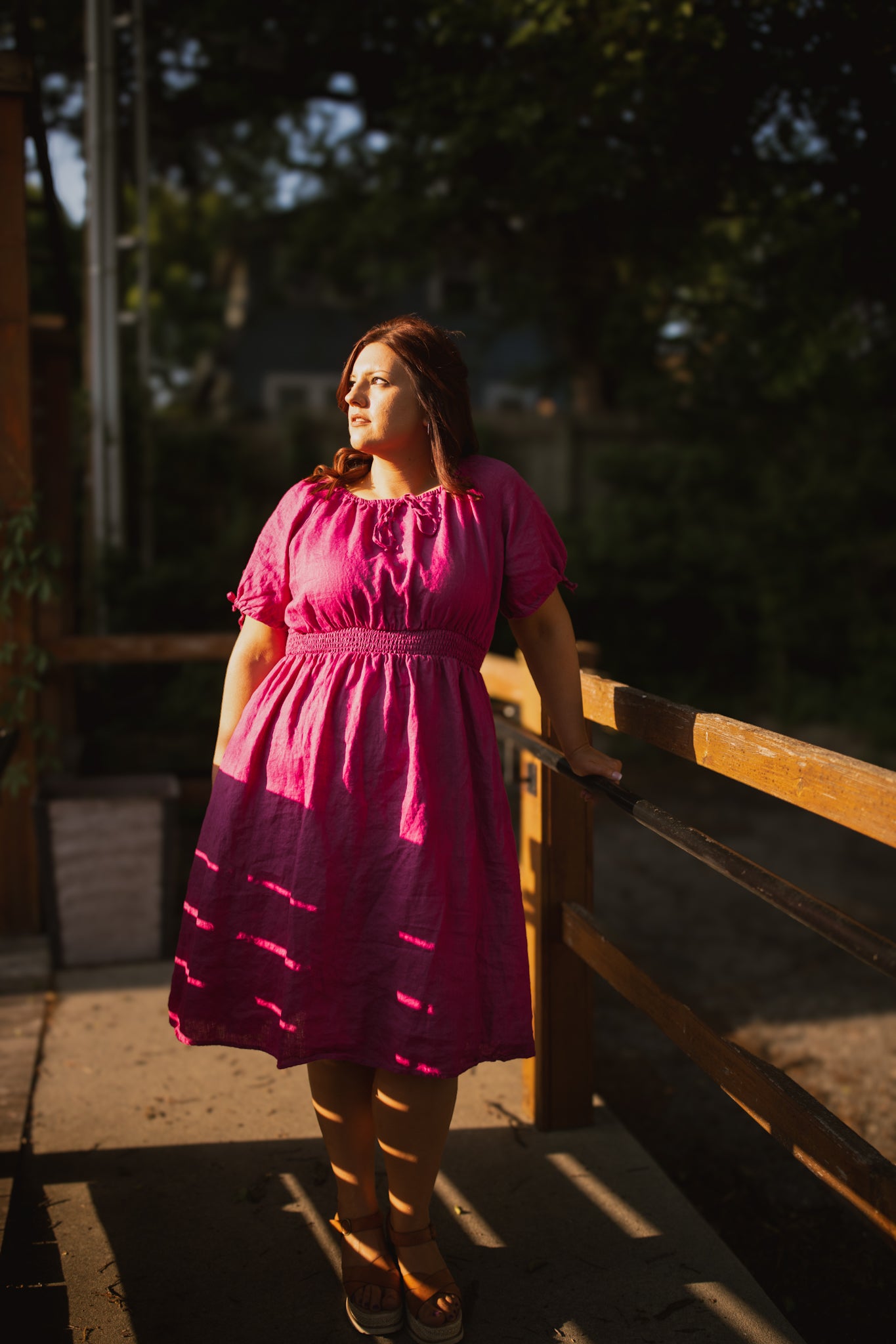 Woman in a pink modest nursing dress standing on a wooden bridge with trees in the background