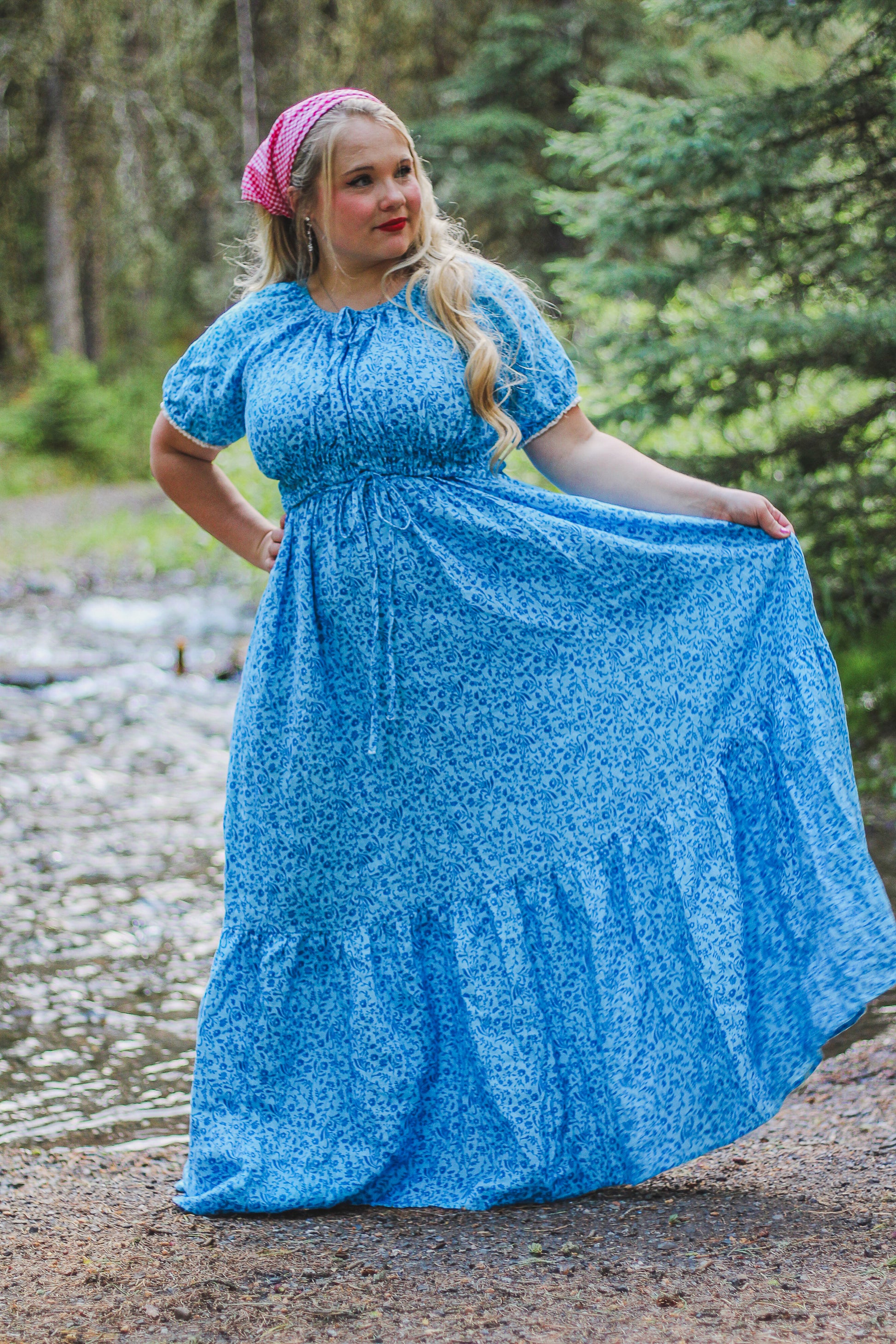 Woman wearing a blue modest nursing dress with puffed sleeves and a pink headscarf, standing outdoors with greenery in the background.