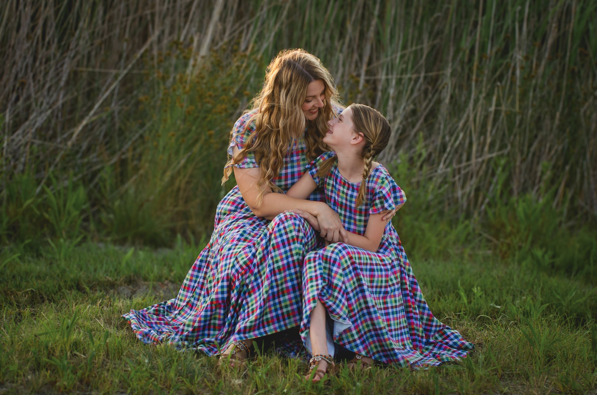 Woman and young girl sitting in a grassy field wearing matching plaid modest dresses.