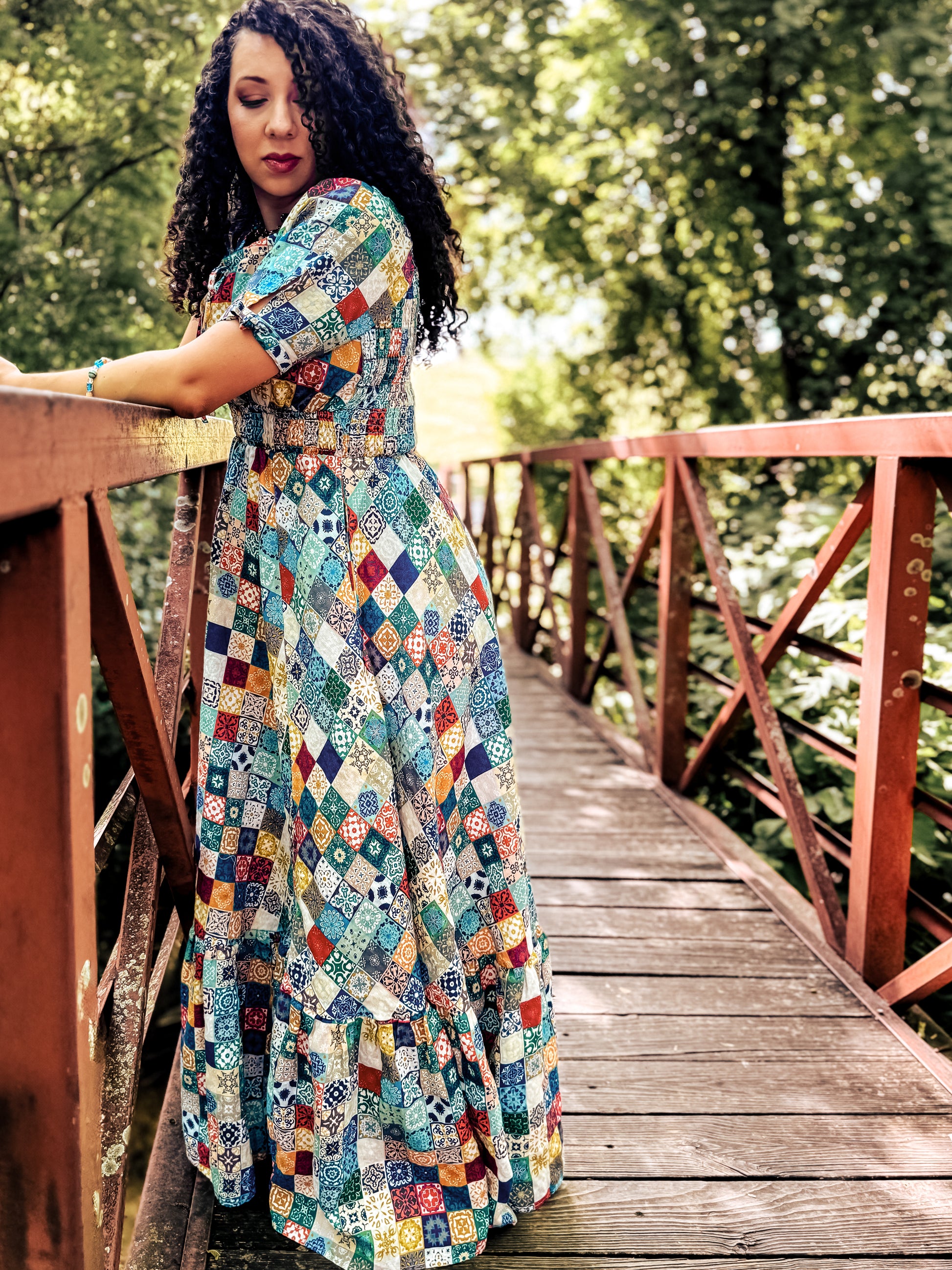 Woman in a colorful patterned modest nursing dress standing on a wooden bridge with greenery in the background