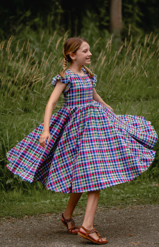 Young girl in a colorful checkered modest dress standing in a grassy area.