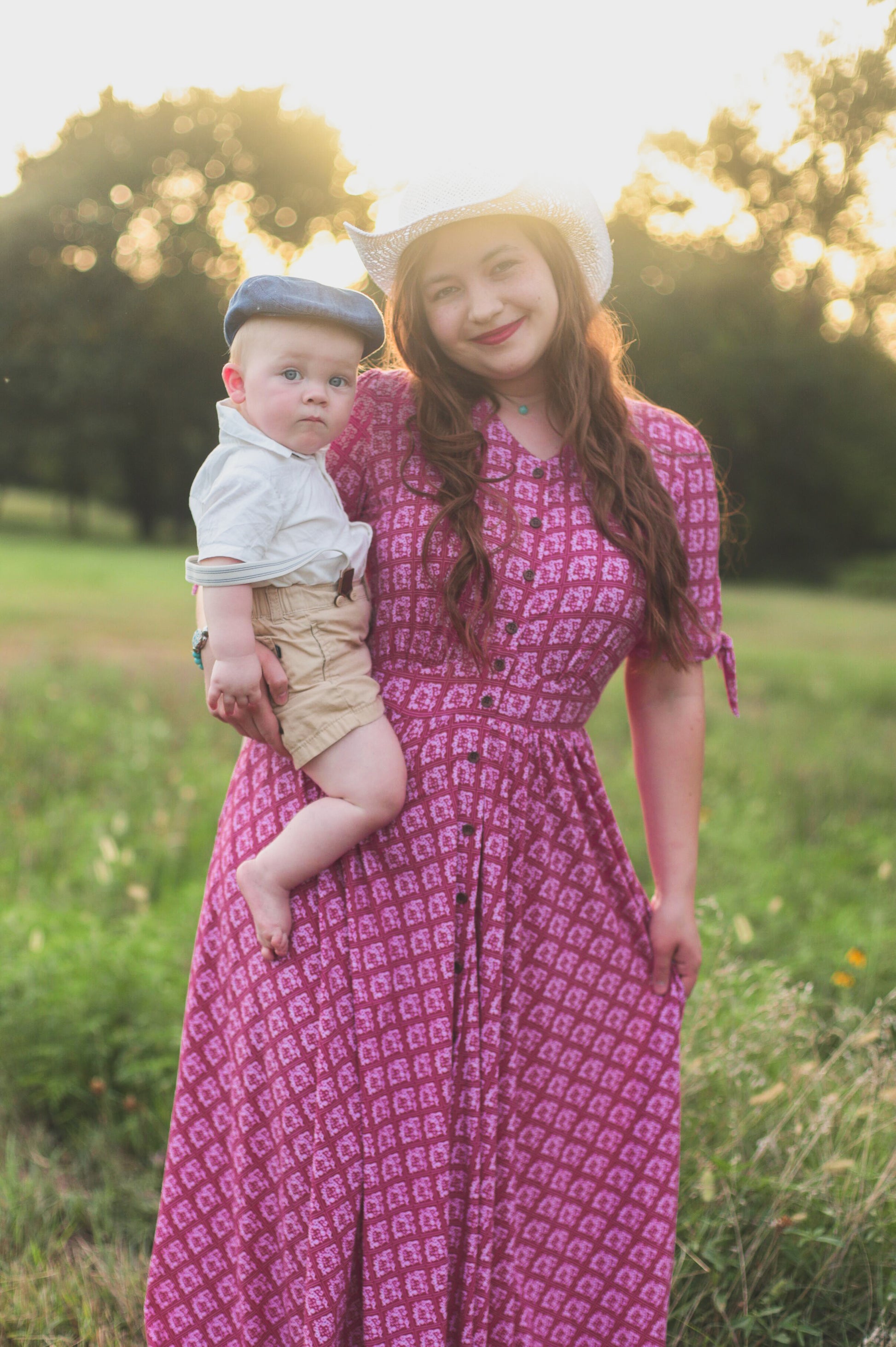 Woman in a pink modest nursing dress holding a child in a field with trees in the background