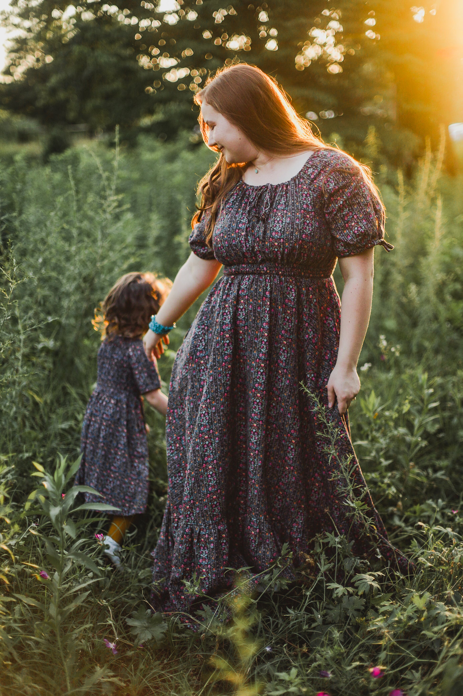 Mother and daughter in floral dresses standing in a field with sunlight filtering through.