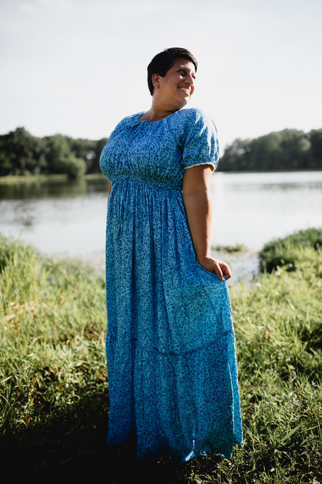 Woman in a blue modest nursing dress standing in a grassy area near a body of water.