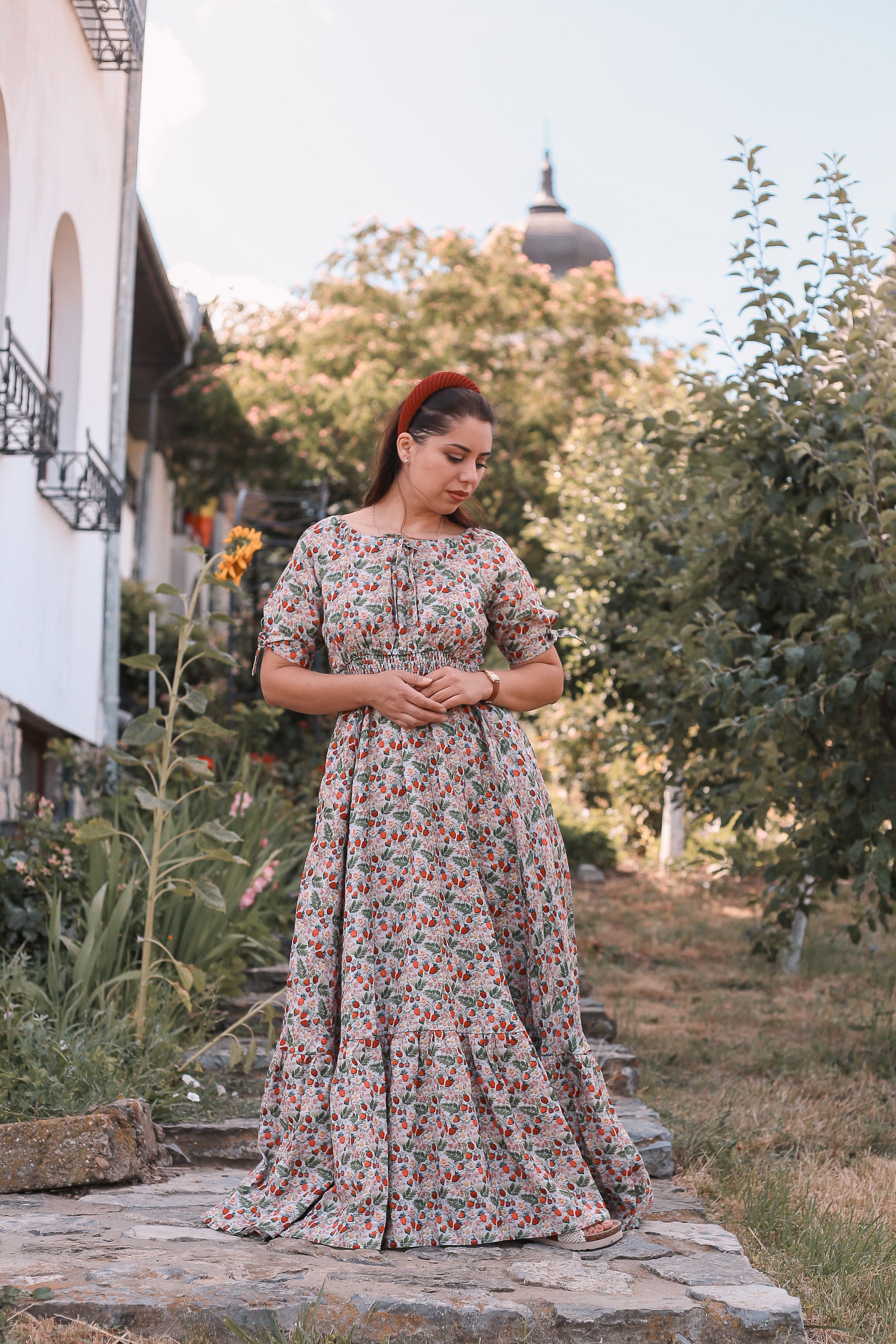 Woman in a floral modest nursing dress standing outdoors with trees and a building in the background