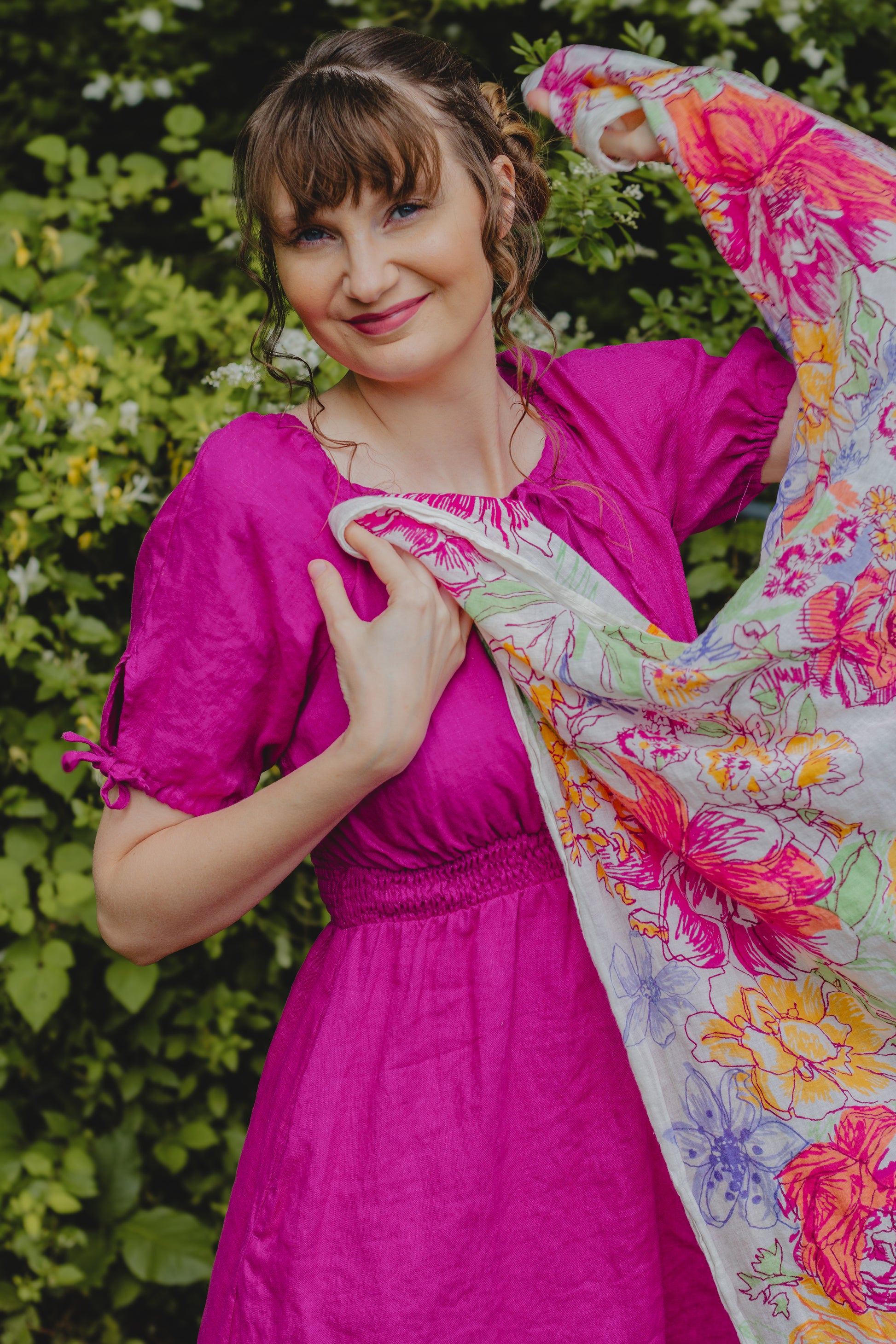 Woman in a pink modest nursing dress holding a colorful floral scarf against a green leafy background