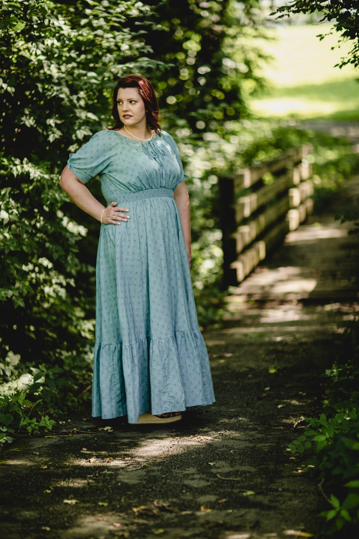 Woman in a blue modest nursing dress standing on a path surrounded by greenery