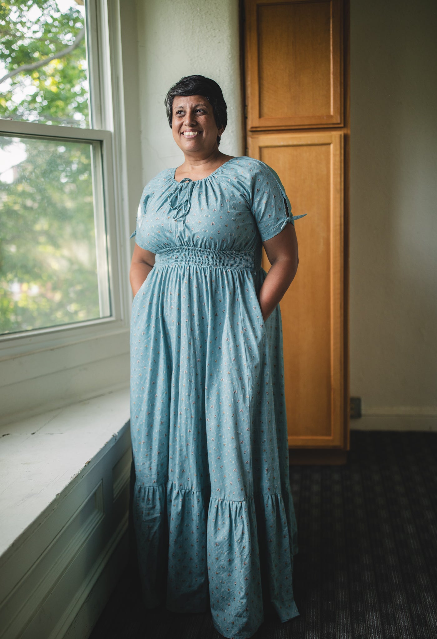 Woman in a blue modest nursing dress standing by a window in a room with wooden cabinets.