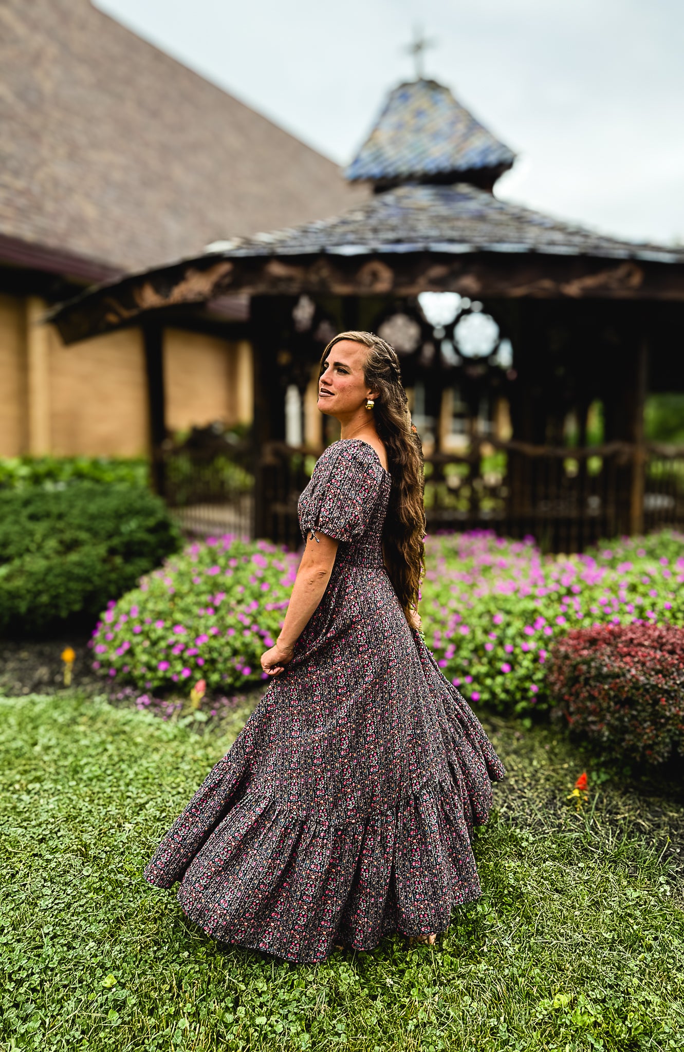 Woman in a floral modest nursing dress standing in a garden with a gazebo in the background