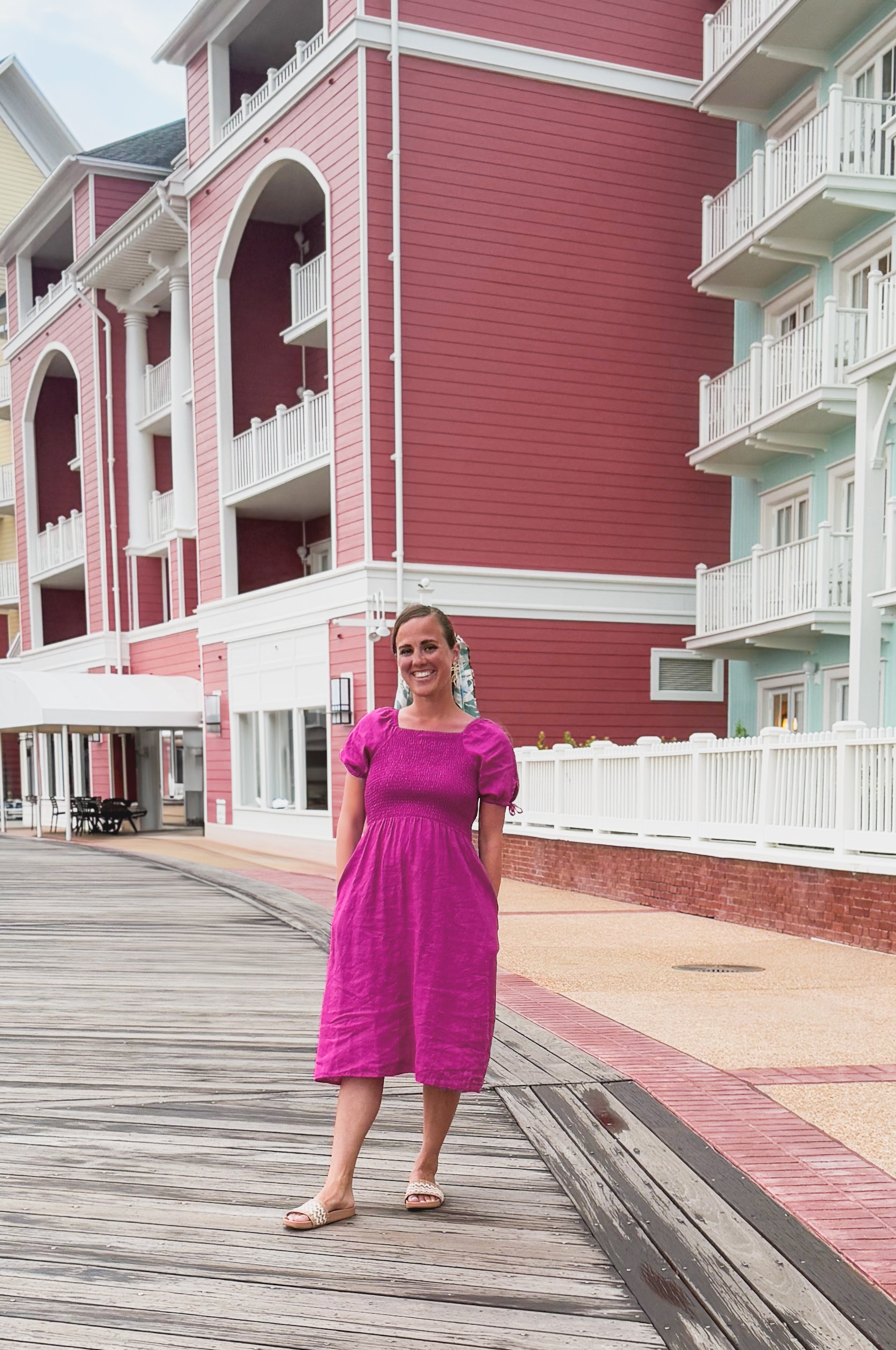 Woman in a pink modest nursing dress standing on a wooden boardwalk in front of a colorful building.