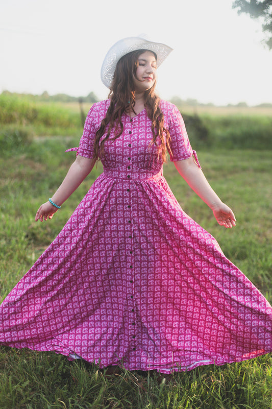 Woman in a pink modest nursing dress and white hat standing in a grassy field