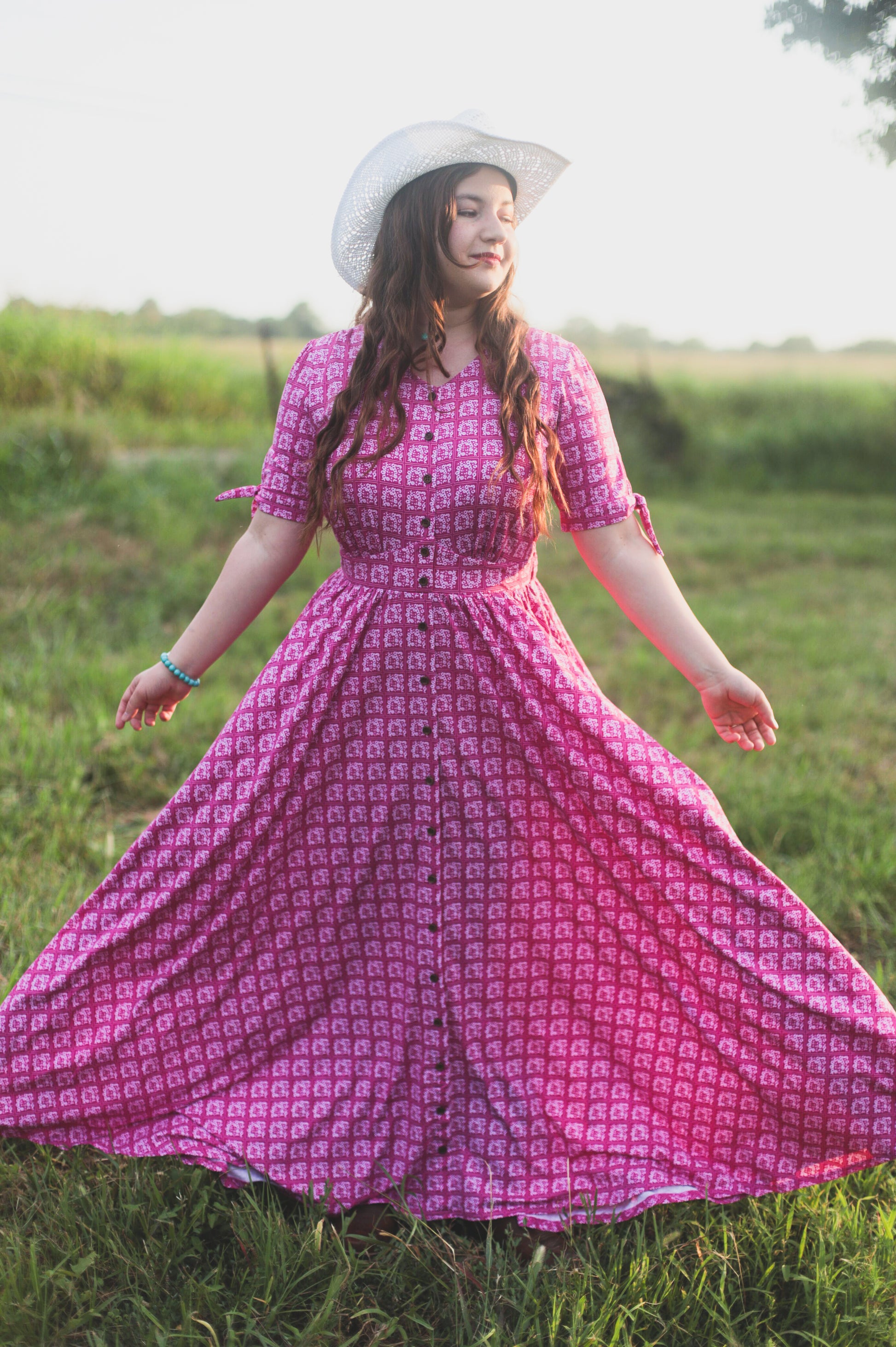 Woman in a pink modest nursing dress and white hat standing in a grassy field