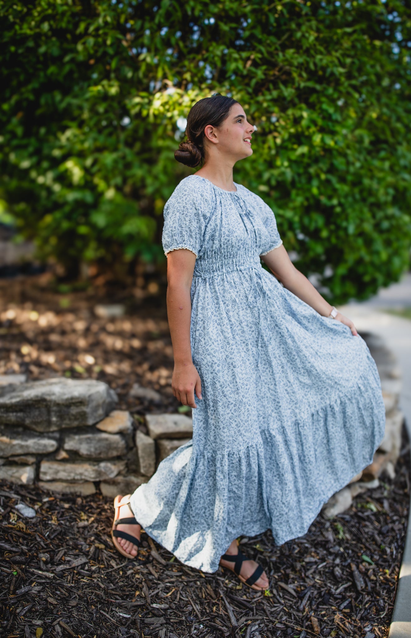 Woman in a modest blue nursing dress standing outdoors with greenery in the background