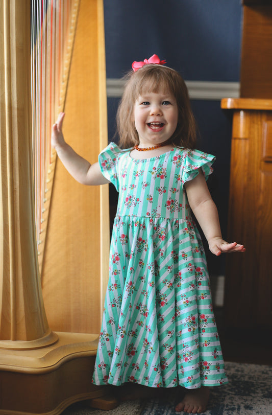 Young girl in a floral modest dress standing in a room with wooden furniture and a blue wall.