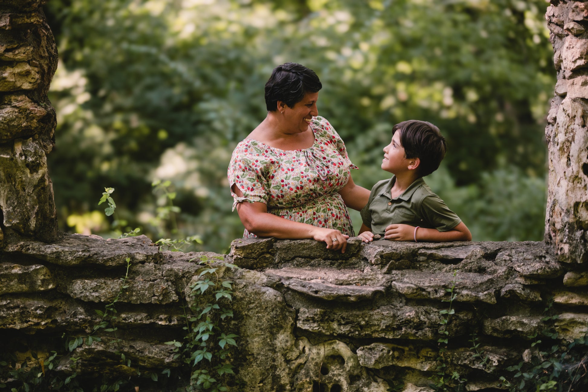 Mother wearing a modest nursing dress and son standing on a stone wall with greenery in the background