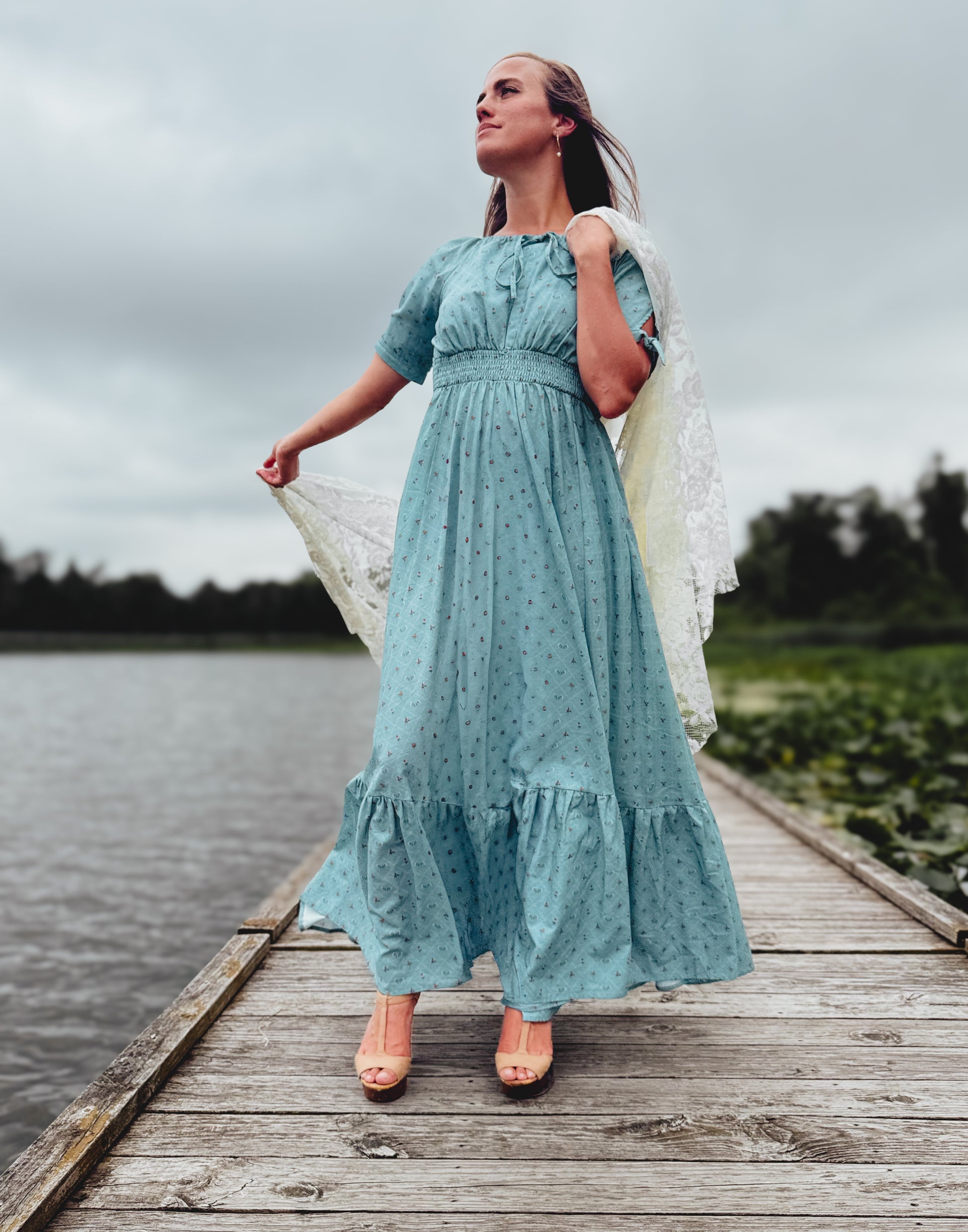 Woman in a light blue modest nursing dress standing on a wooden dock by a lake.