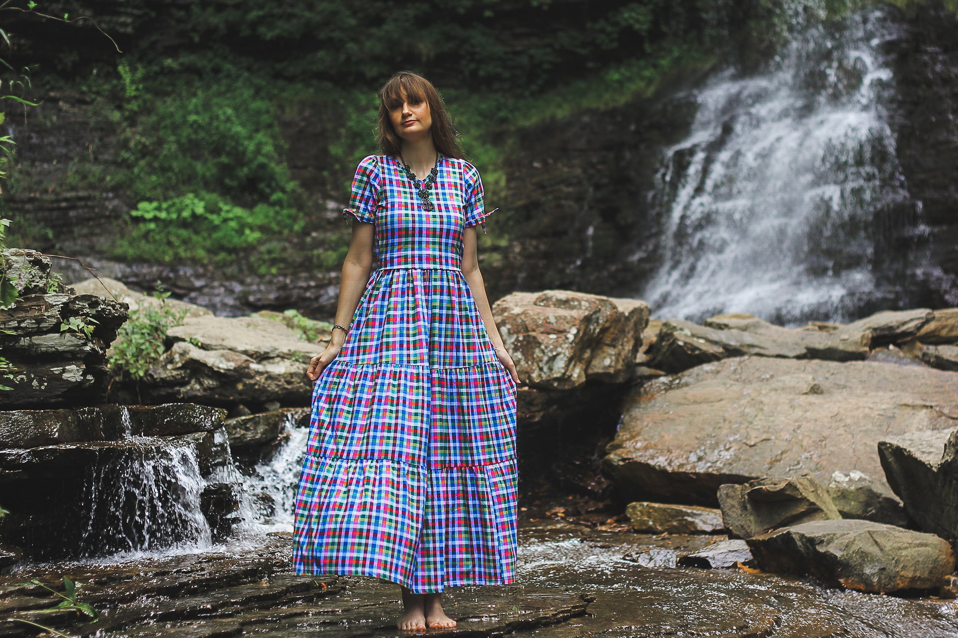 Woman in a plaid modest nursing dress standing in front of a waterfall