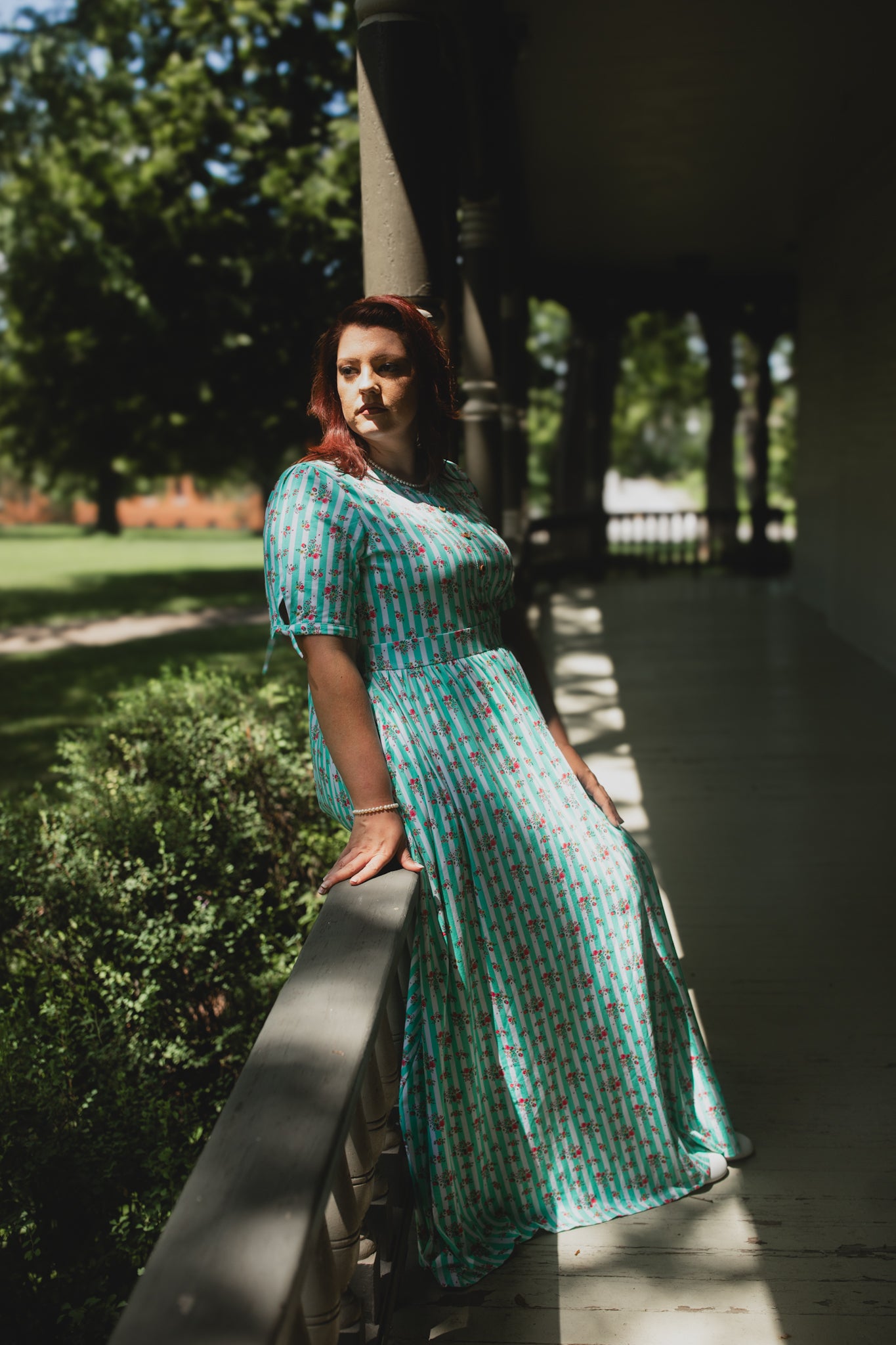 Woman in a green patterned modest nursing dress standing outdoors with trees and a building in the background