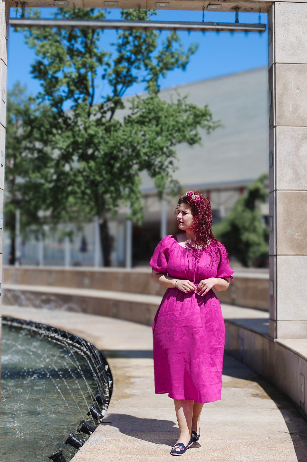 Woman in a pink modest nursing dress standing near a water fountain with trees and a building in the background.
