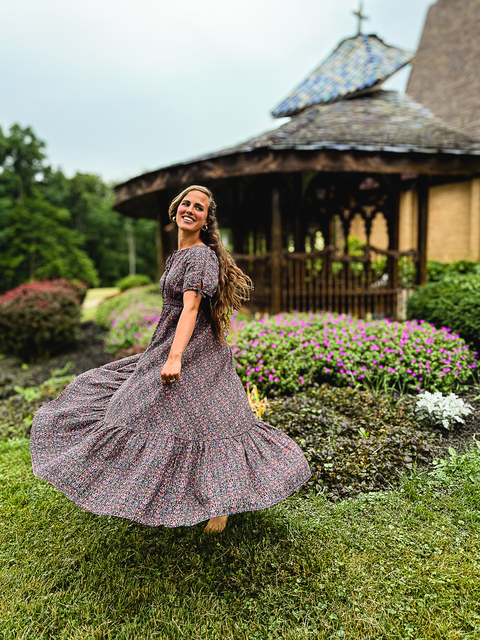 Woman in a floral modest nursing dress standing in a garden with a gazebo in the background