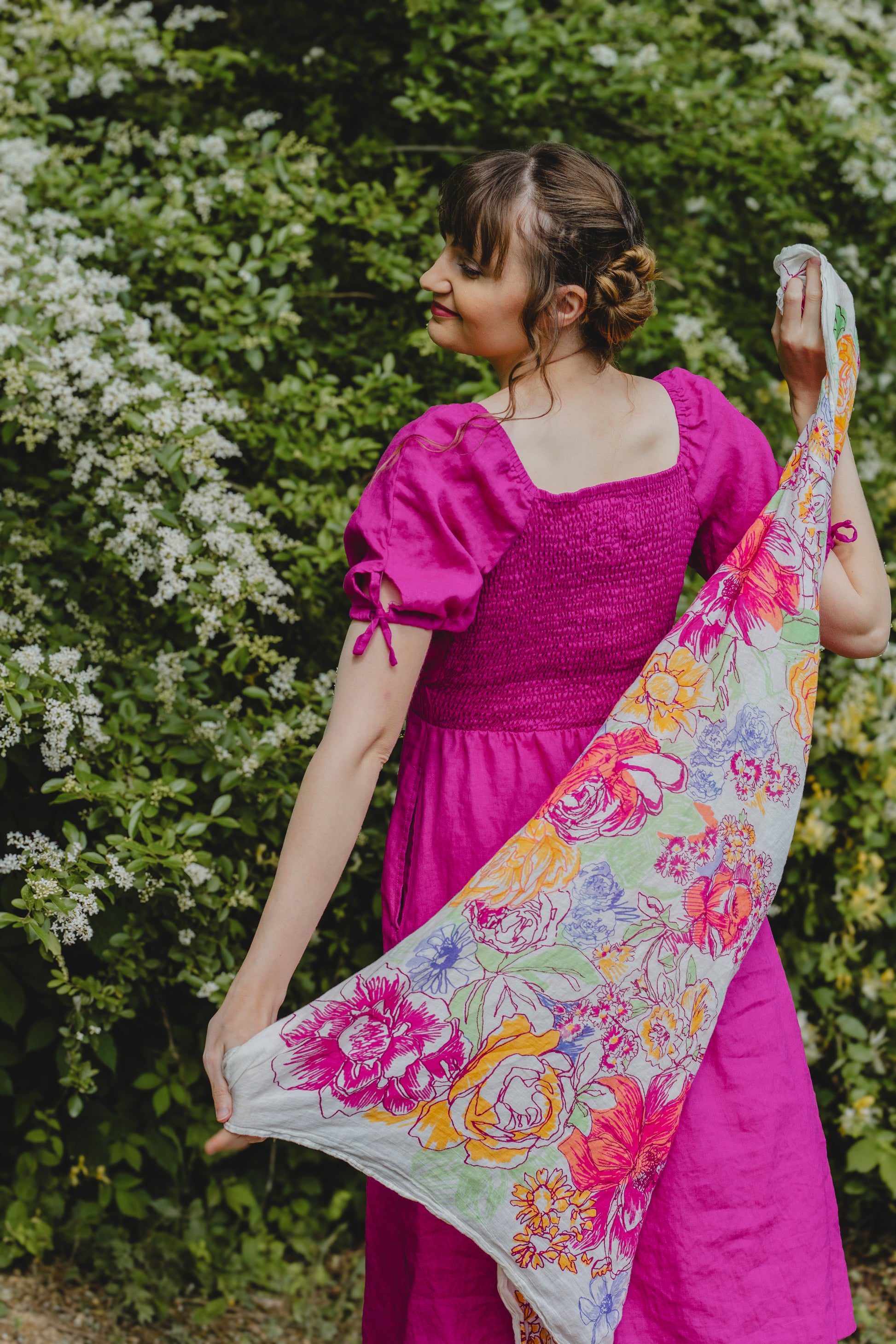 Woman in a pink modest nursing dress holding a colorful floral scarf against a green bush background