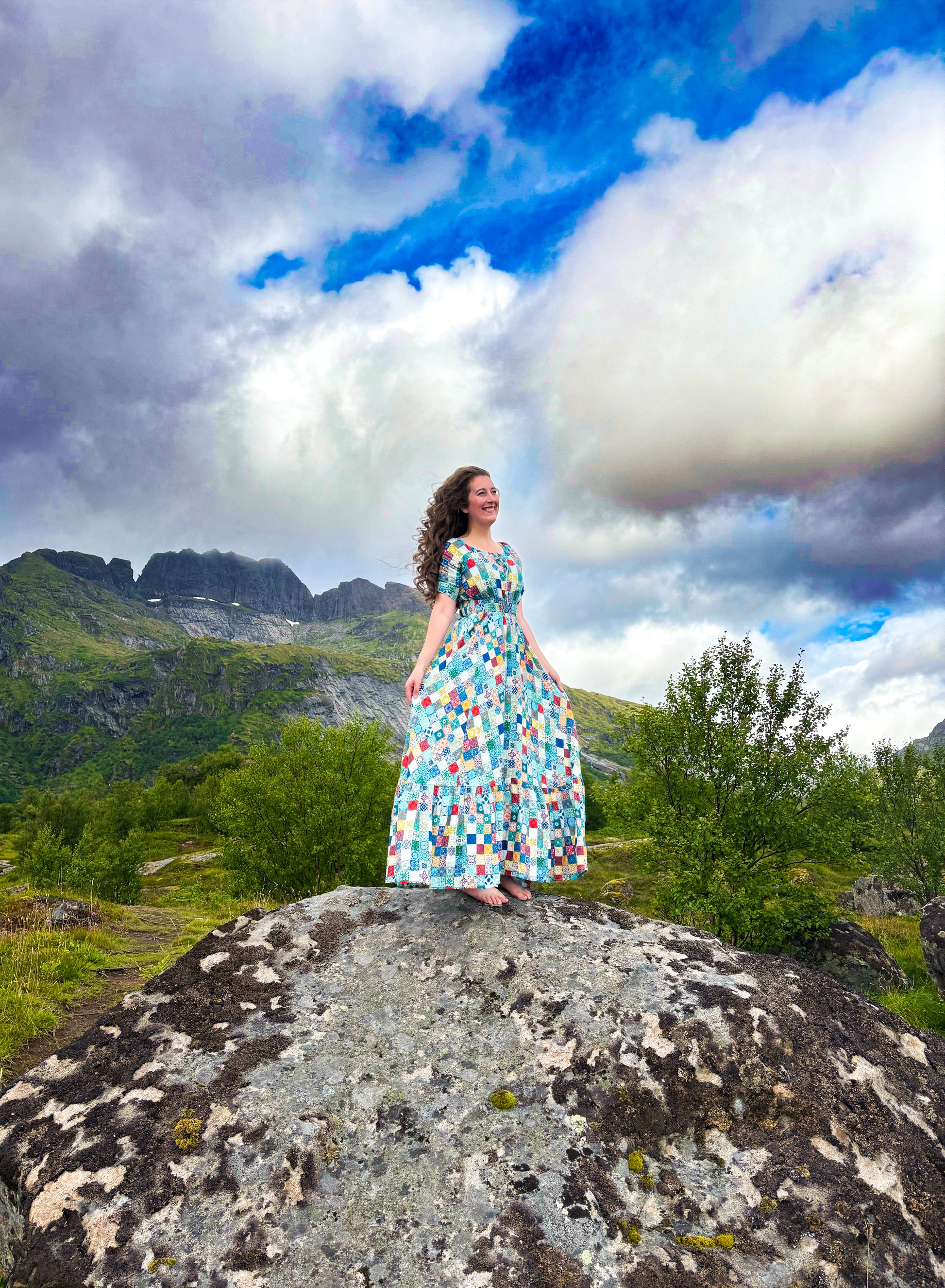 Woman in a colorful modest nursing dress standing on a rock with mountains and clouds in the background