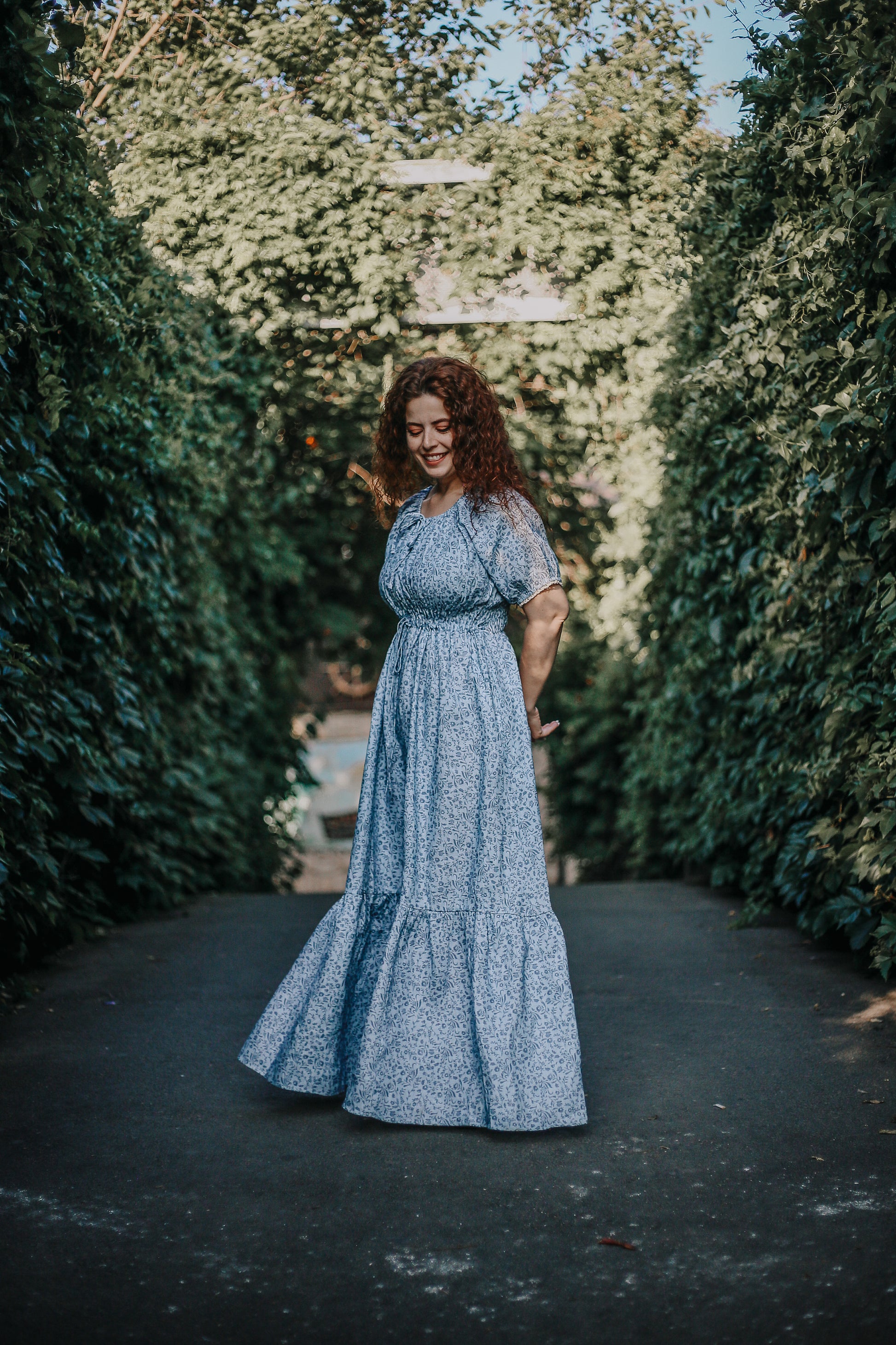 Woman in a modest blue nursing dress standing in a garden path lined with green hedges.