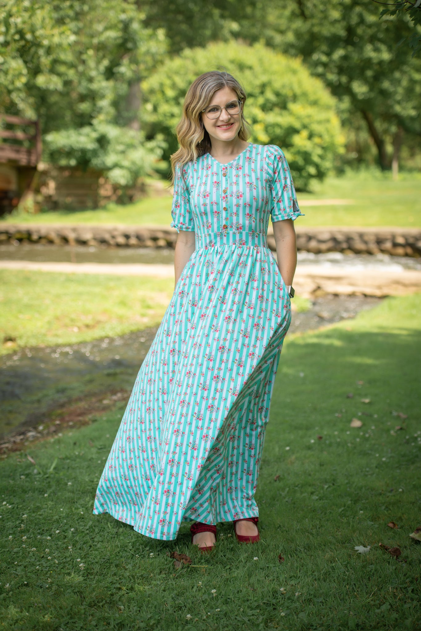 Woman in a long, patterned modest nursing dress standing outdoors on grass with trees in the background