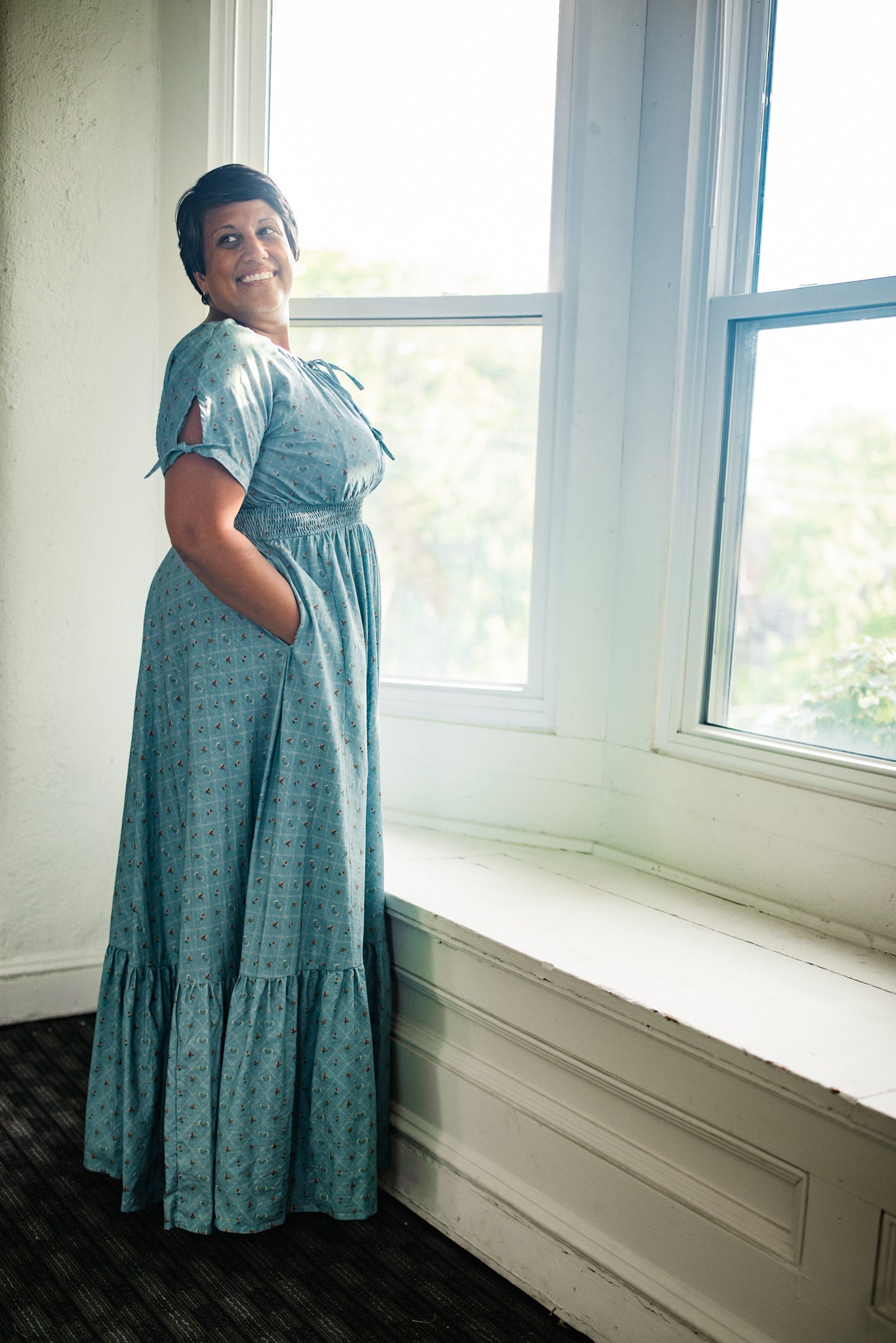 Woman in a teal modest nursing dress standing by a window in a well-lit room.