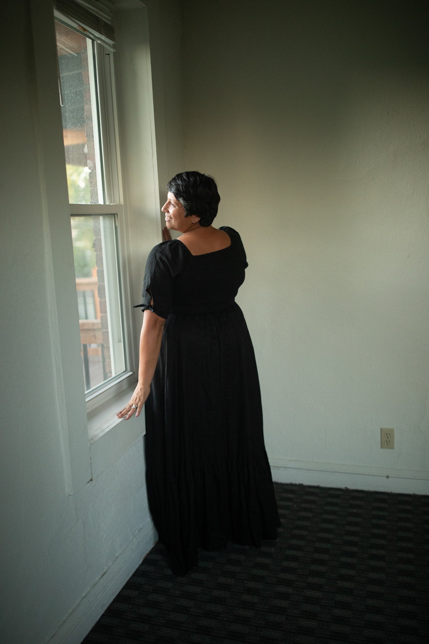 Woman in a black modest nursing dress standing by a window in a room with plain walls.