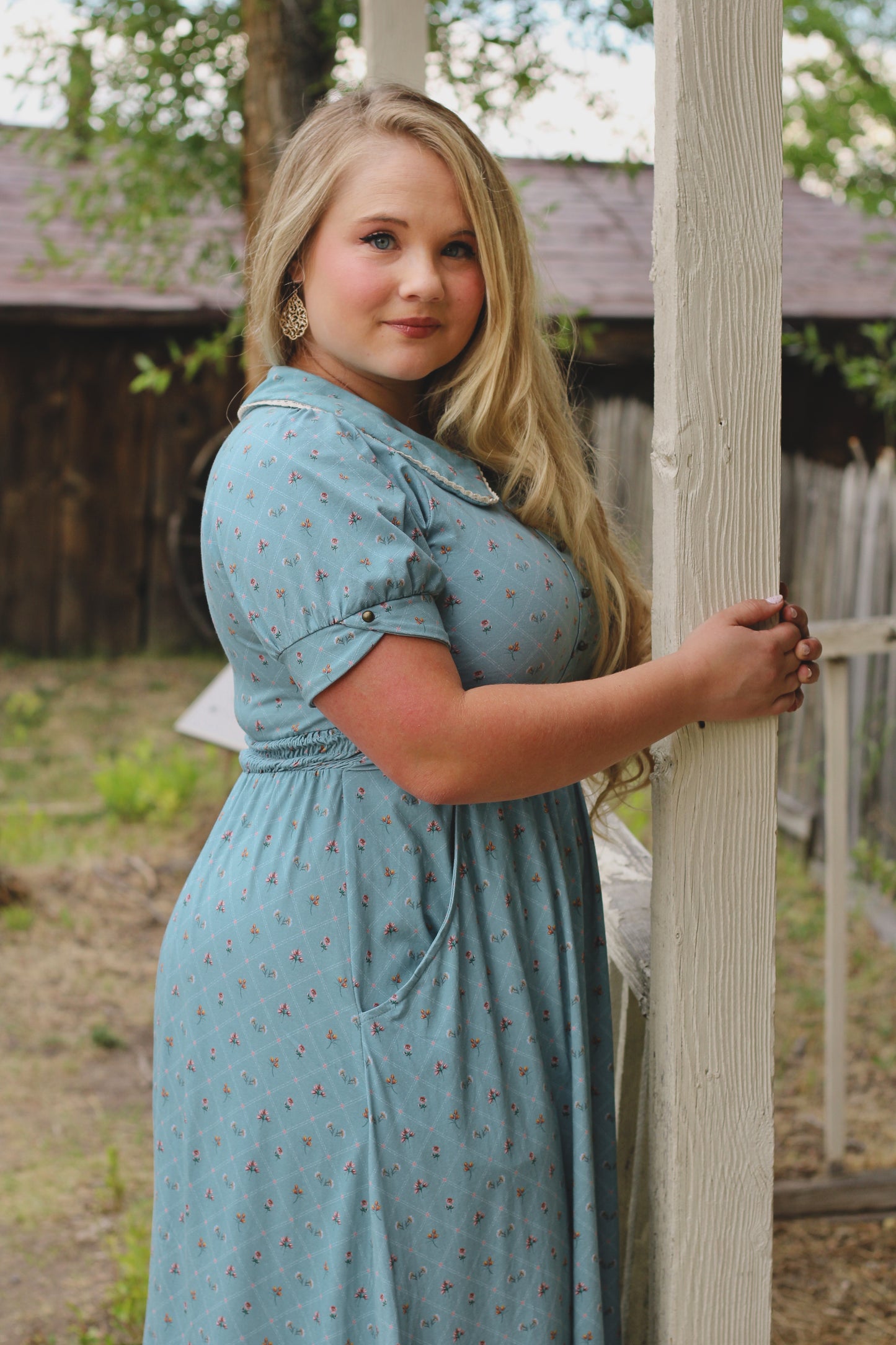Woman in a modest nursing dress standing outdoors near a wooden post.