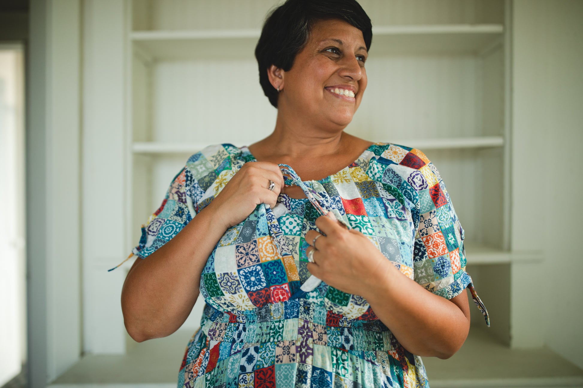 Woman wearing a colorful patchwork modest nursing dress standing in a room with shelves.