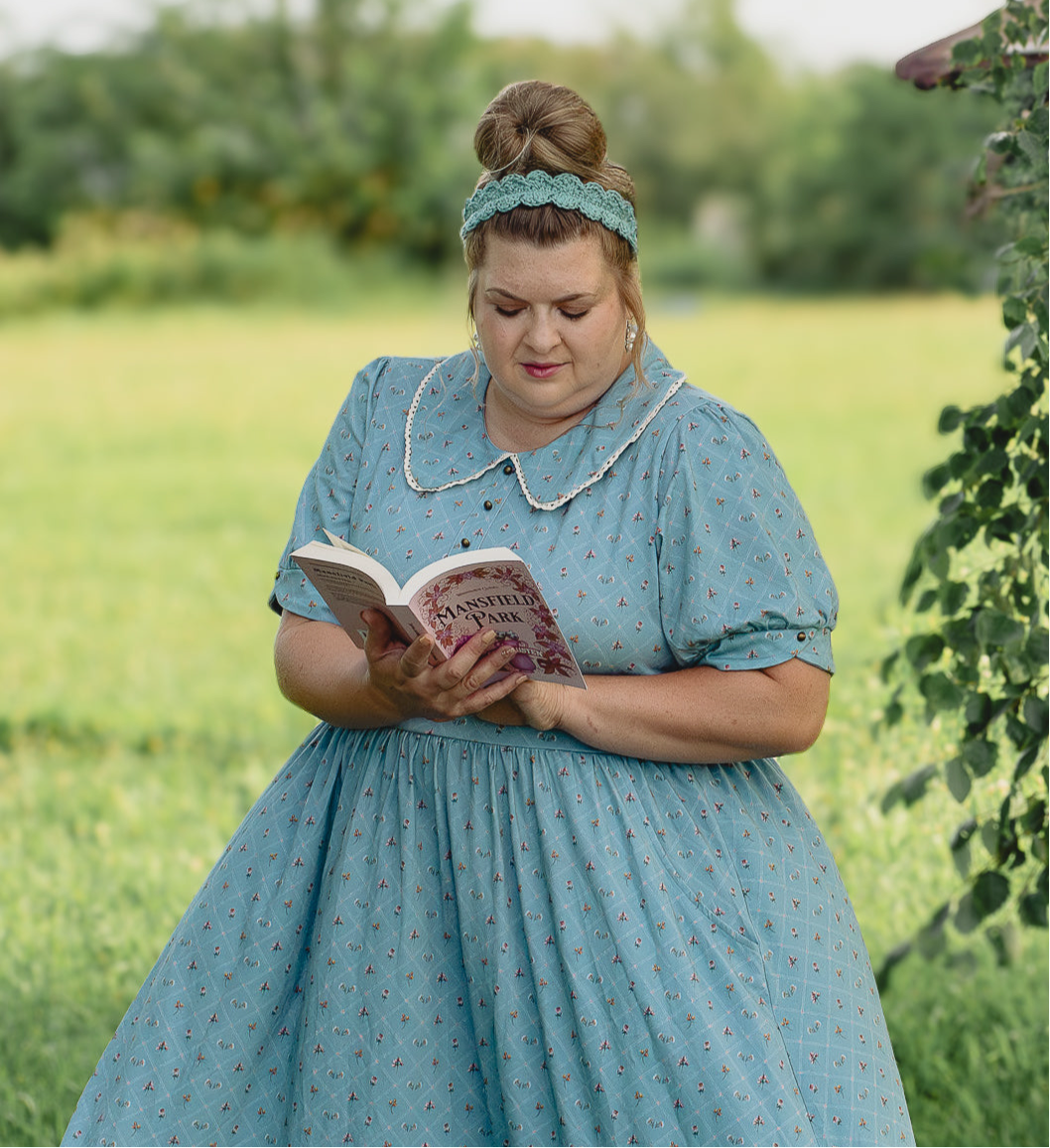 Woman in a modest nursing dress reading a book in a green field
