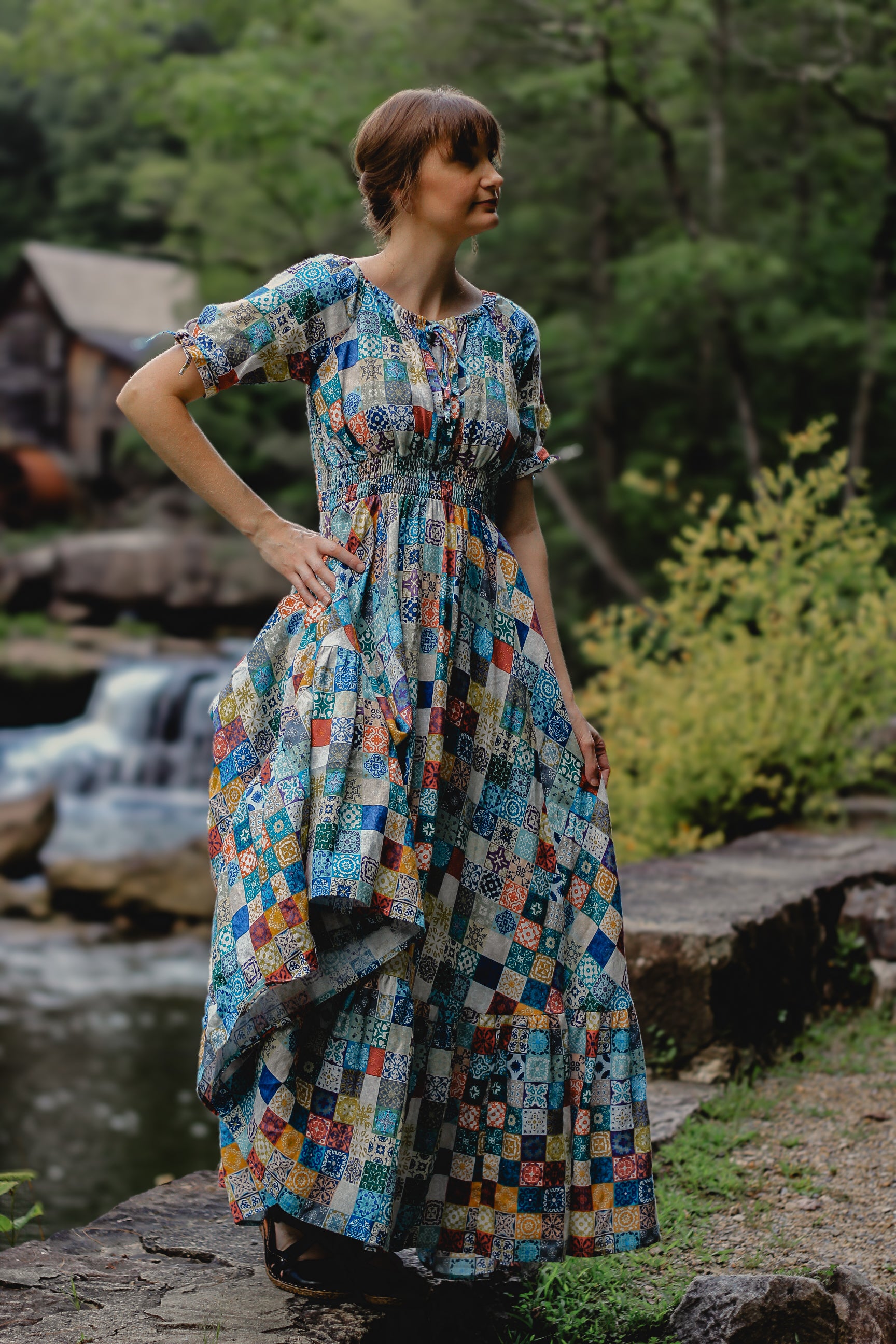 Woman in a colorful modest nursing dress standing by a stream with trees in the background