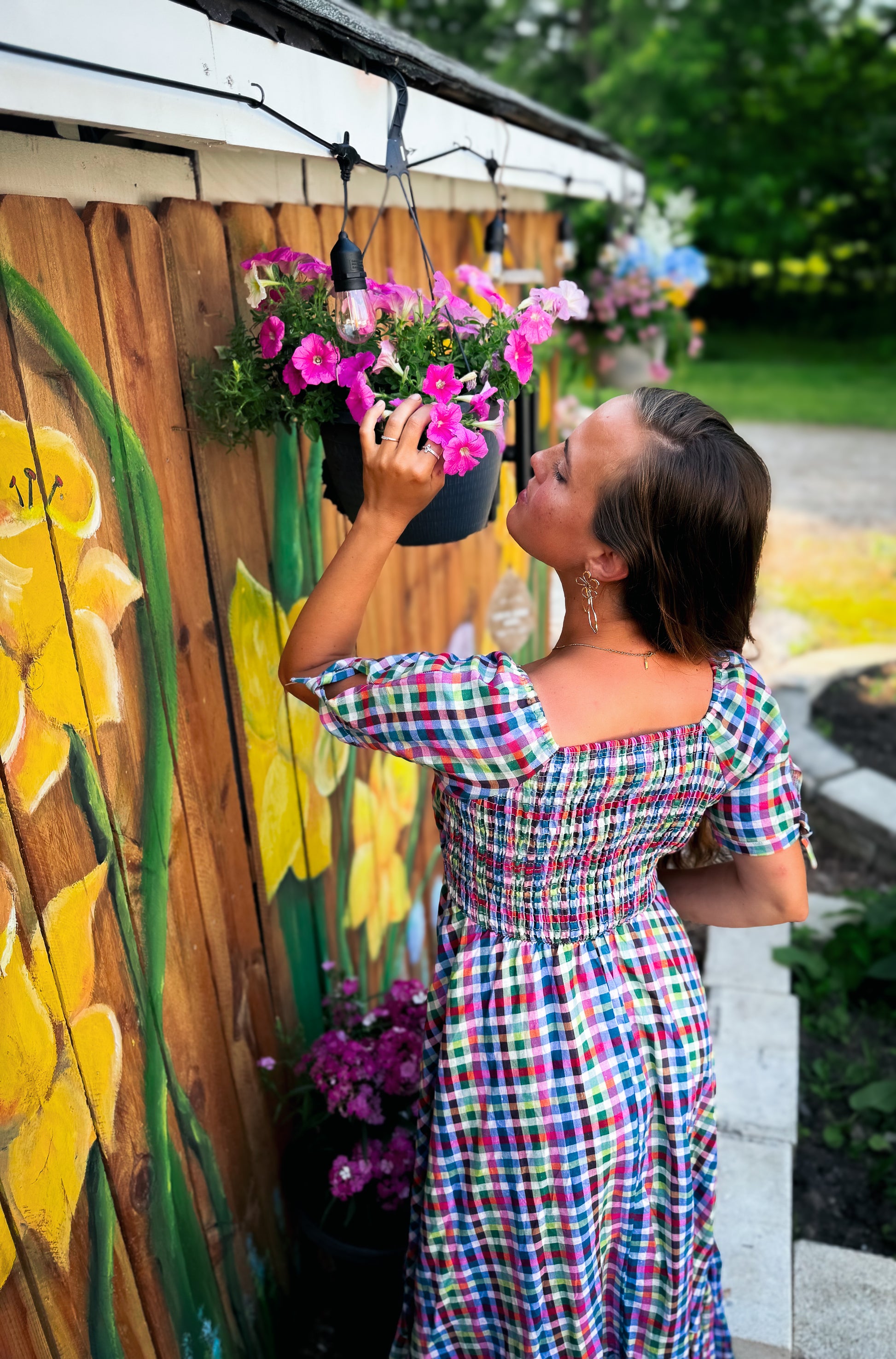 Woman in a colorful modest nursing dress tending to flowers against a wooden fence with floral art.